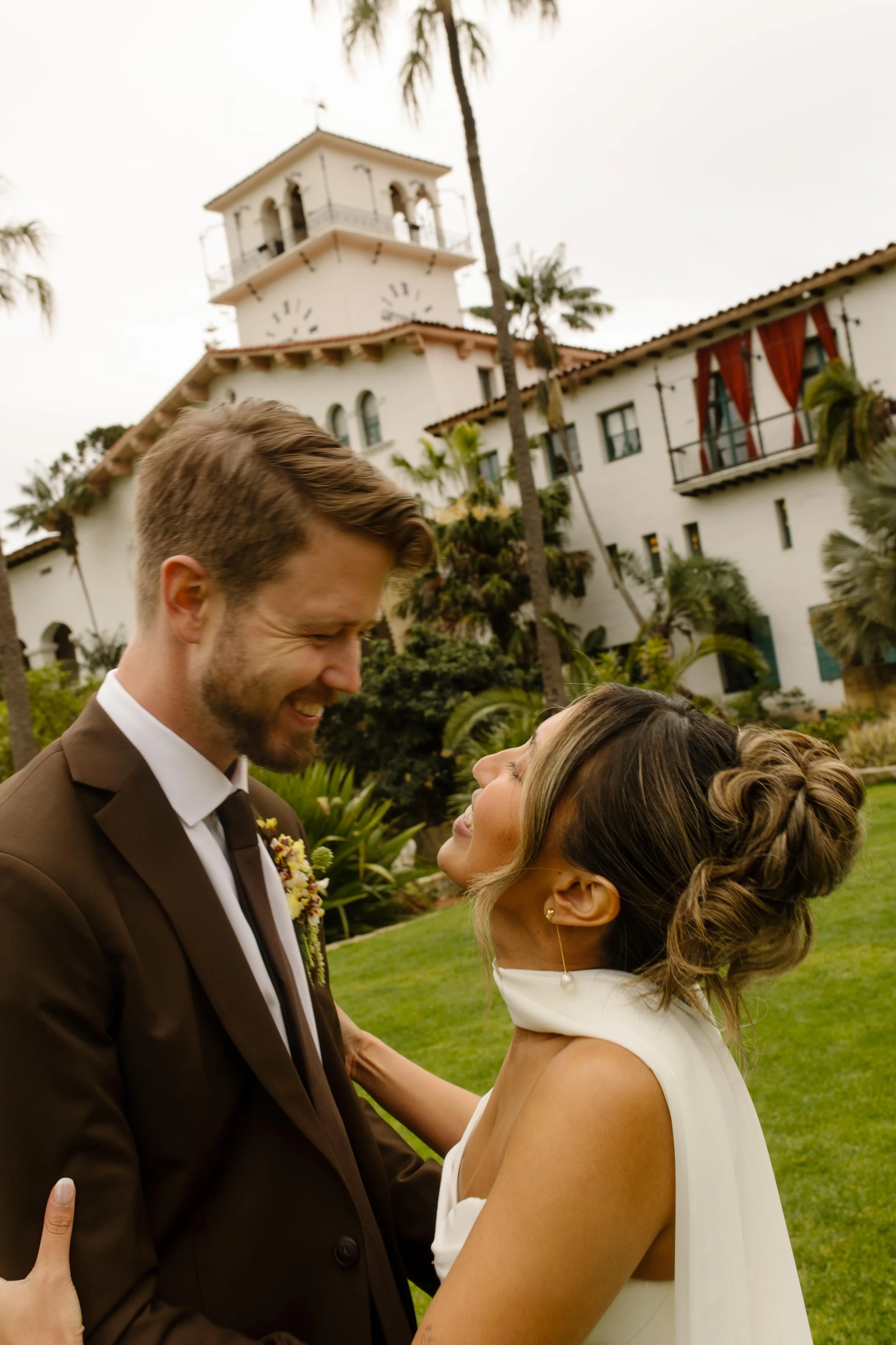 The bride and groom smile at each other in front of the iconic white tower of the Santa Barbara Courthouse, lush palm trees swaying in the background.
