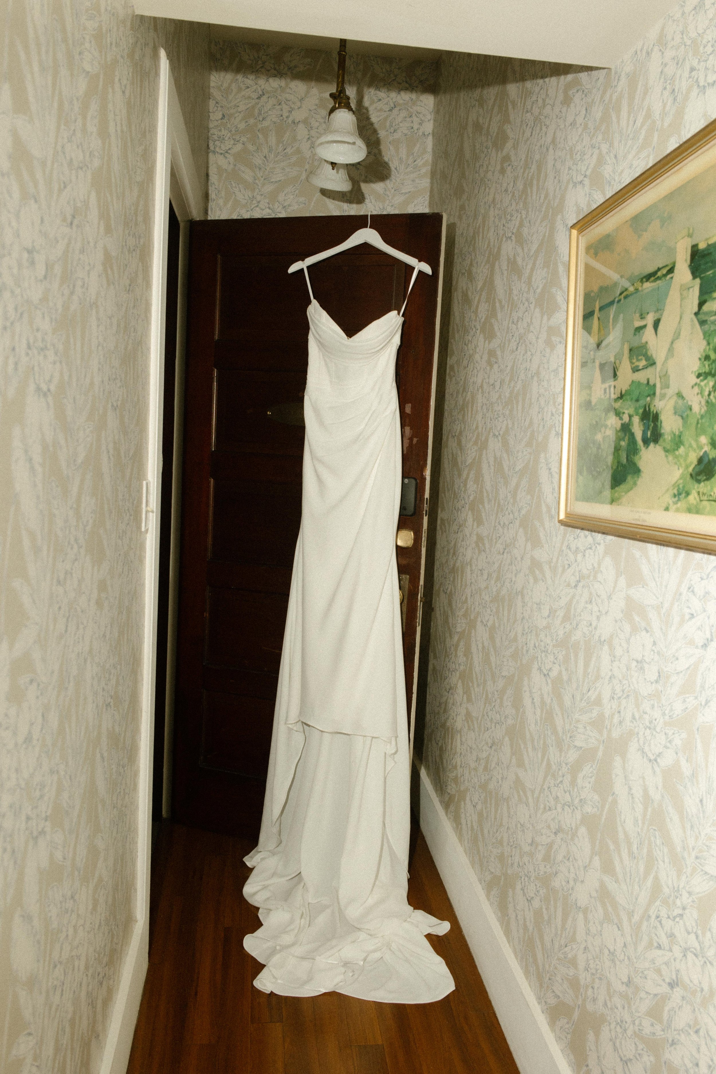 The bride’s gown hangs at the end of a wallpapered hallway, ready and waiting in the calm before the day begins.