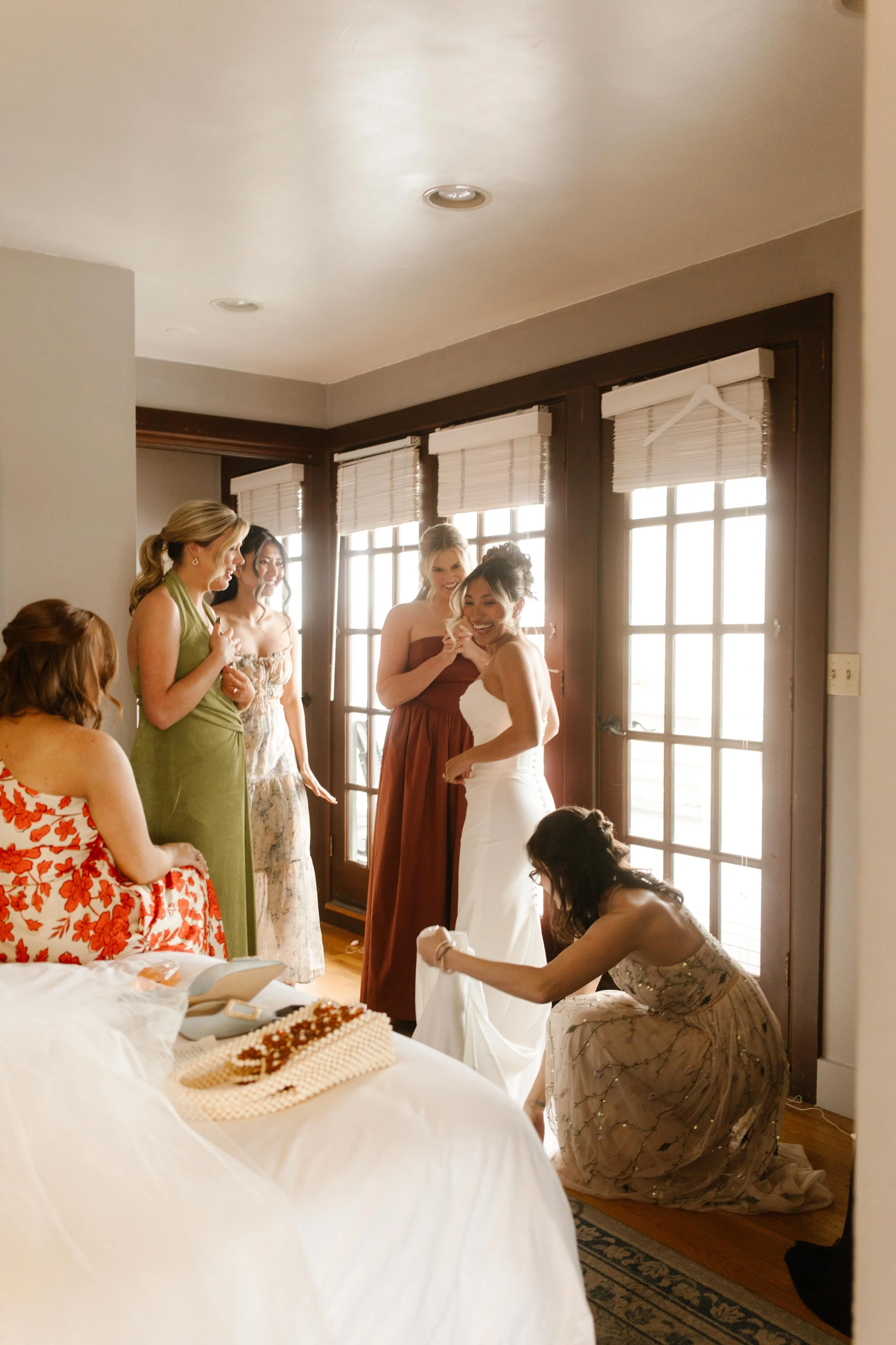 A candid moment as the bride is helped into her dress, surrounded by bridesmaids in colorful dresses, sunlight pouring through the windows.