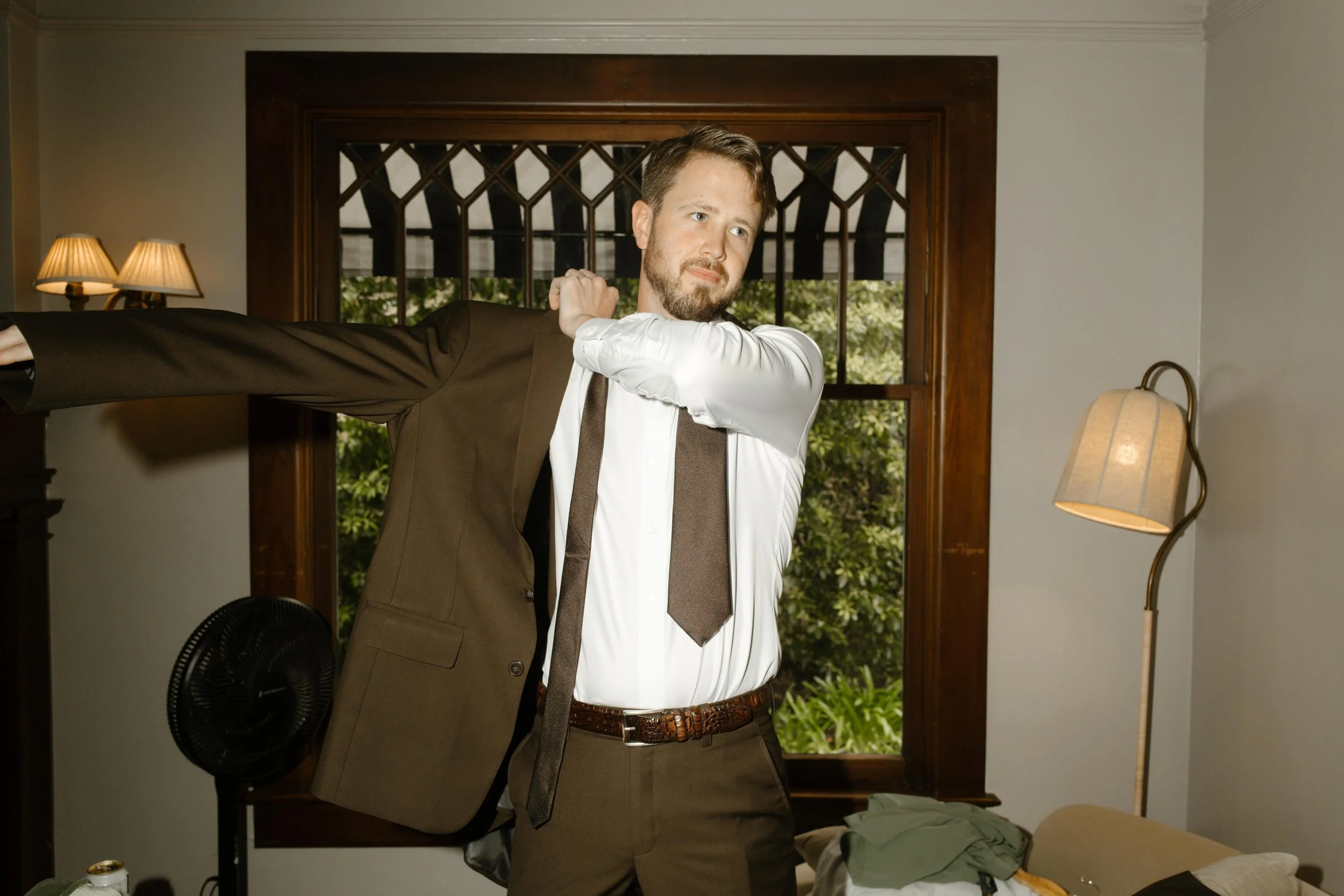 The groom puts on his brown suit jacket, standing by a window filled with soft natural light and leafy green views