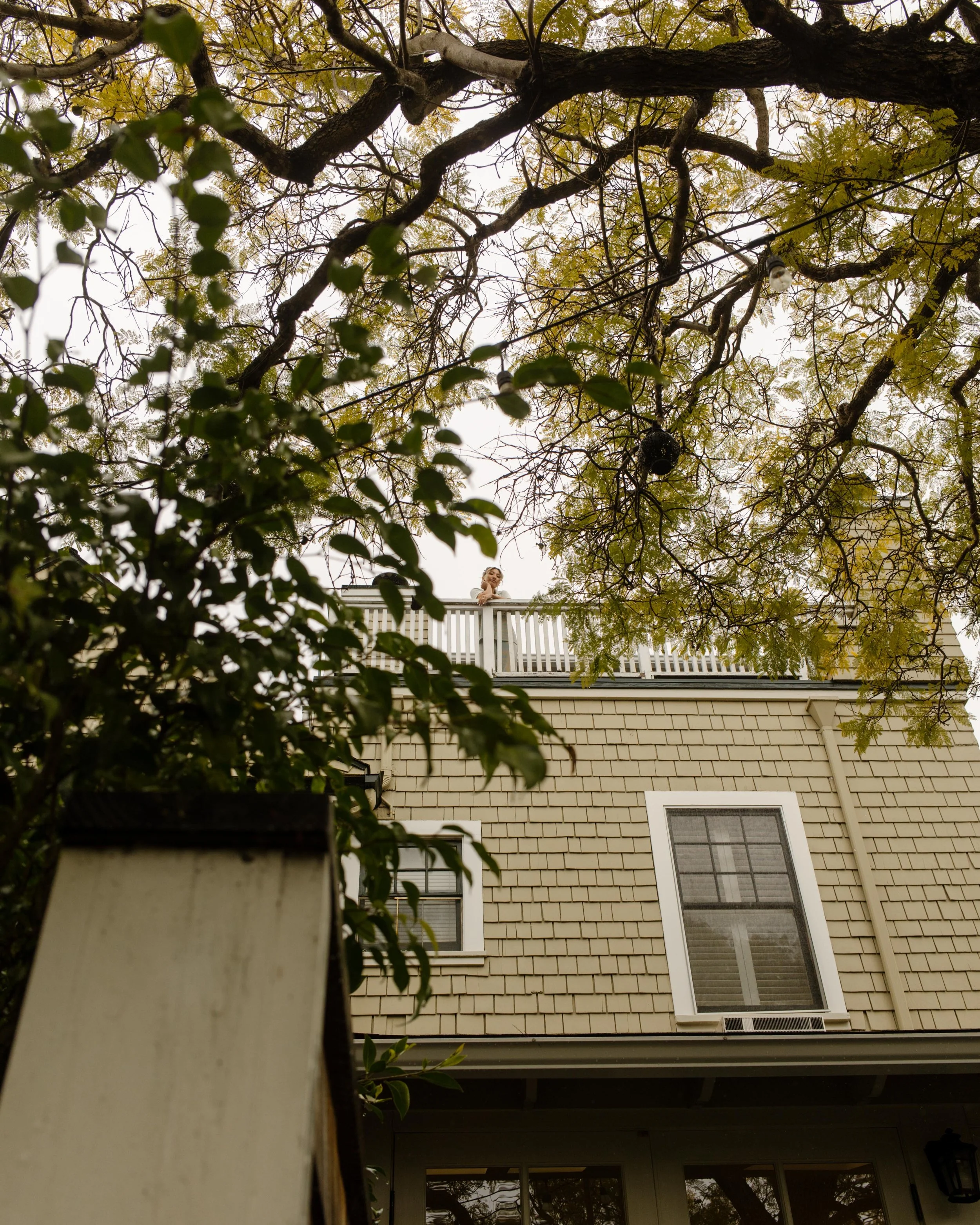 A view from below of the bride standing on a wraparound porch, framed by leafy branches and a gray shingle house.