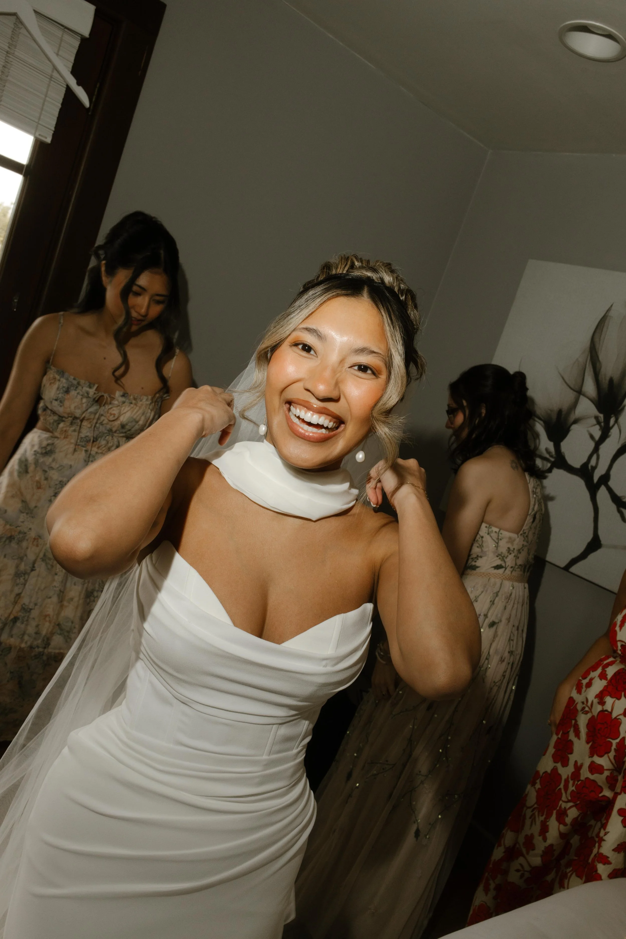 The bride laughs while adjusting her veil, surrounded by bridesmaids in floral dresses and the hum of getting-ready energy.