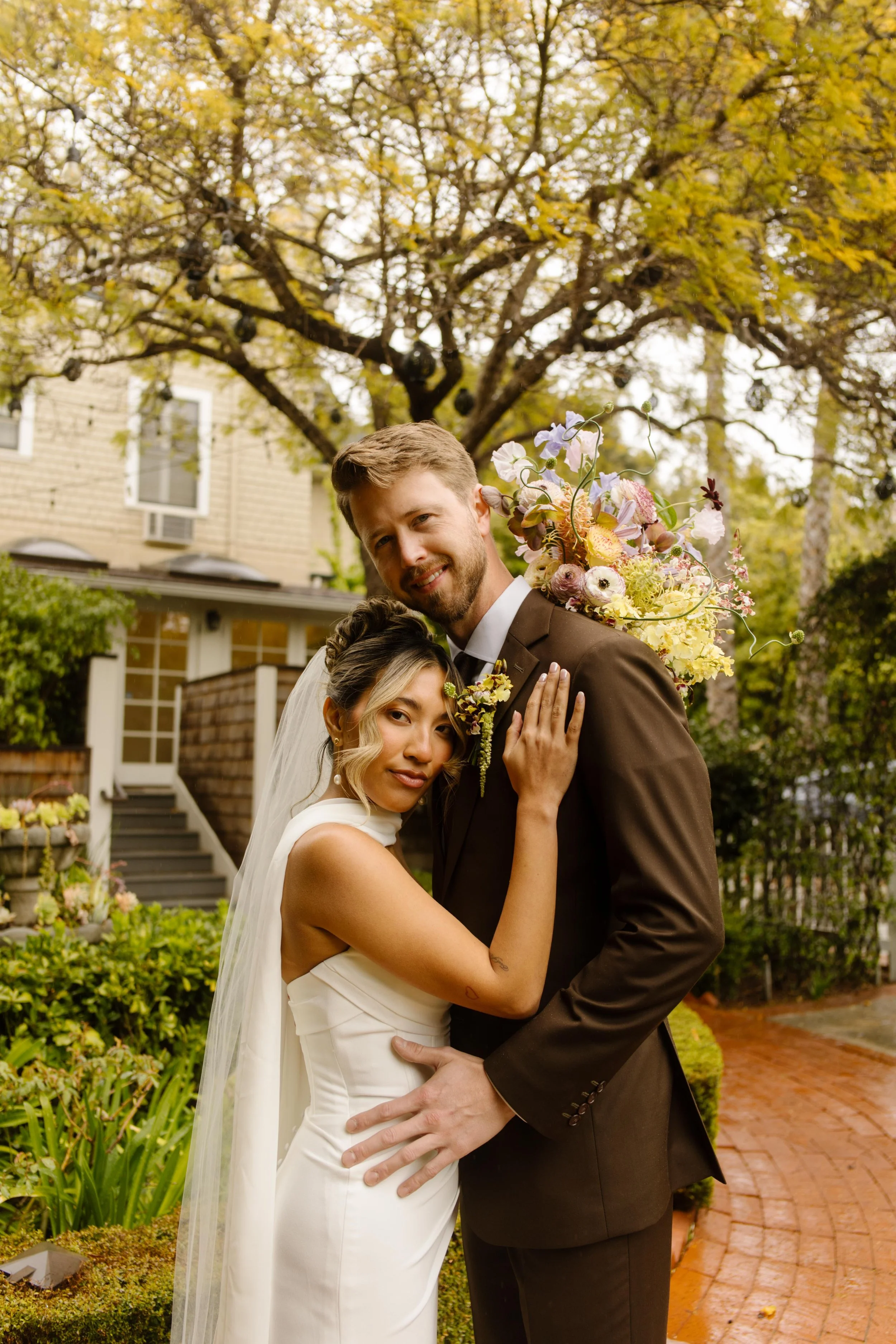 The bride leans into the groom with a quiet smile, framed by golden trees and the soft brick path of their Santa Barbara celebration.