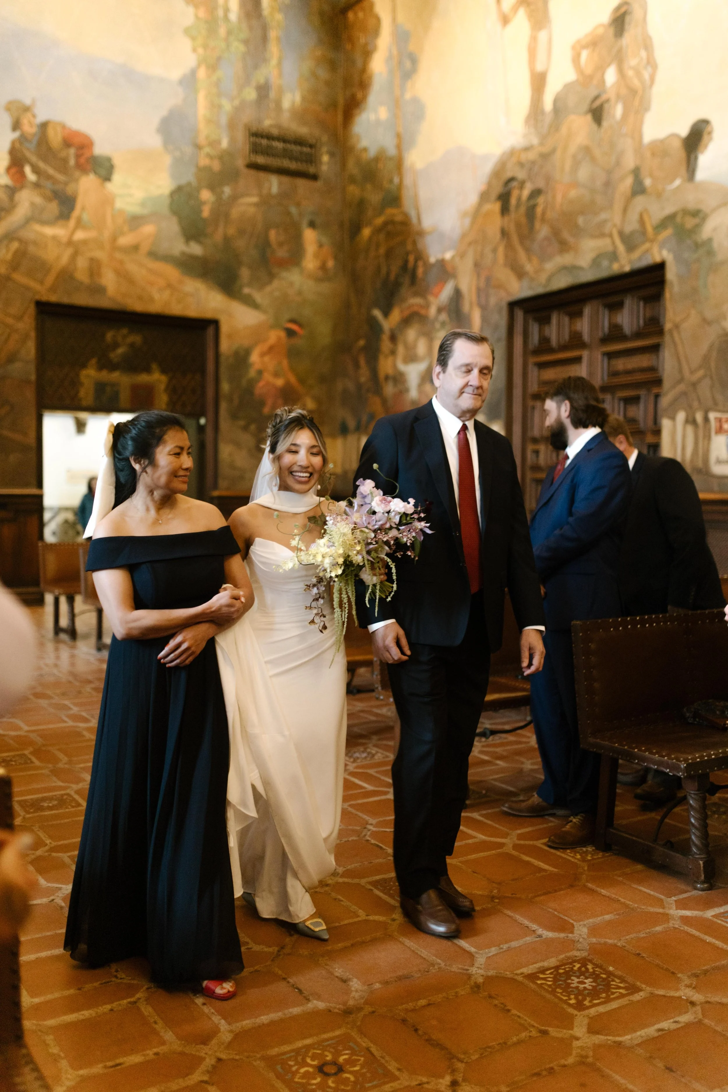 The bride walks down the aisle with her parents, beaming as she enters the mural room surrounded by sweeping frescoes.