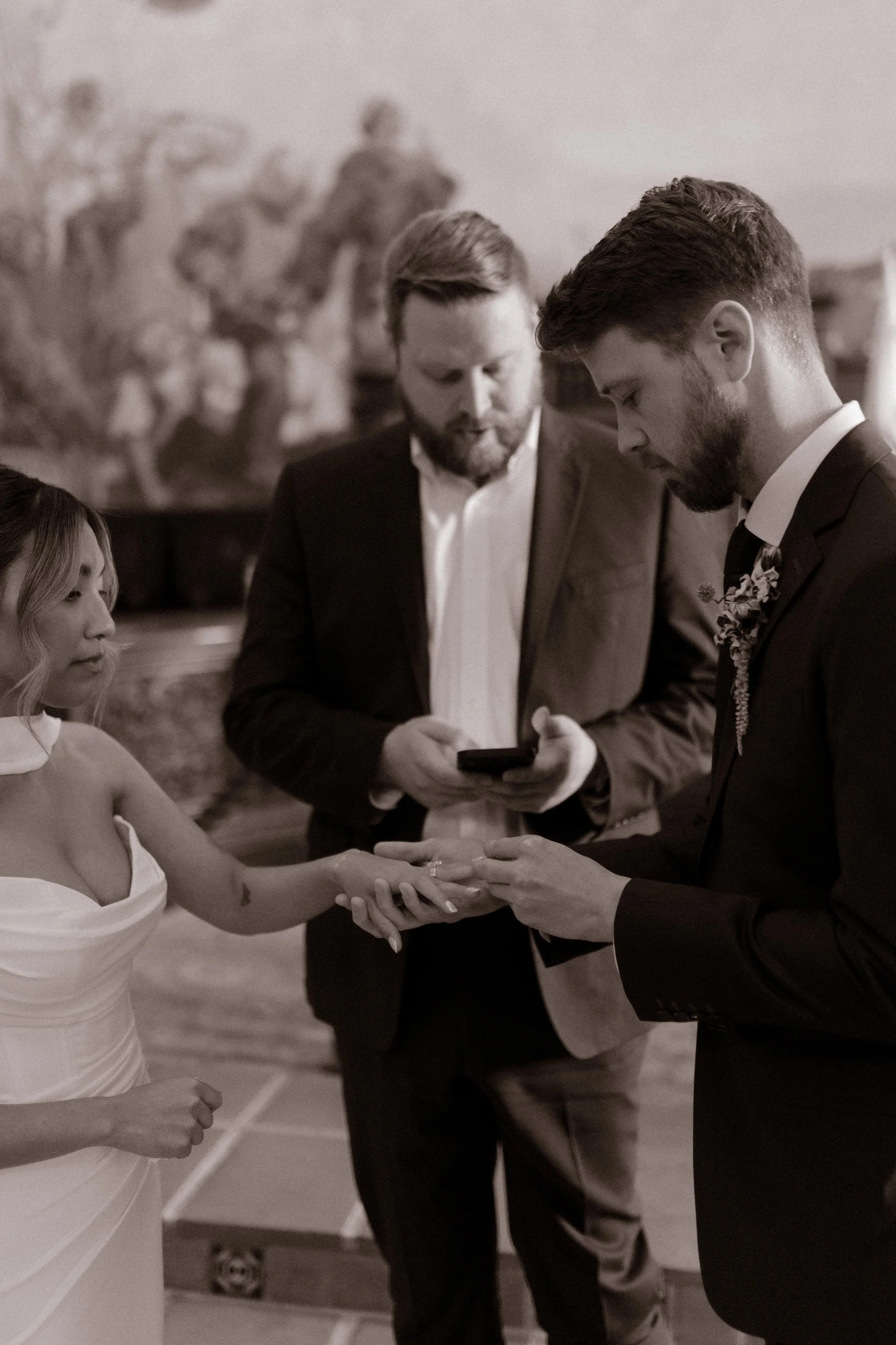 A close-up, sepia-toned moment of the groom placing the ring on the bride’s finger mid-ceremony.
