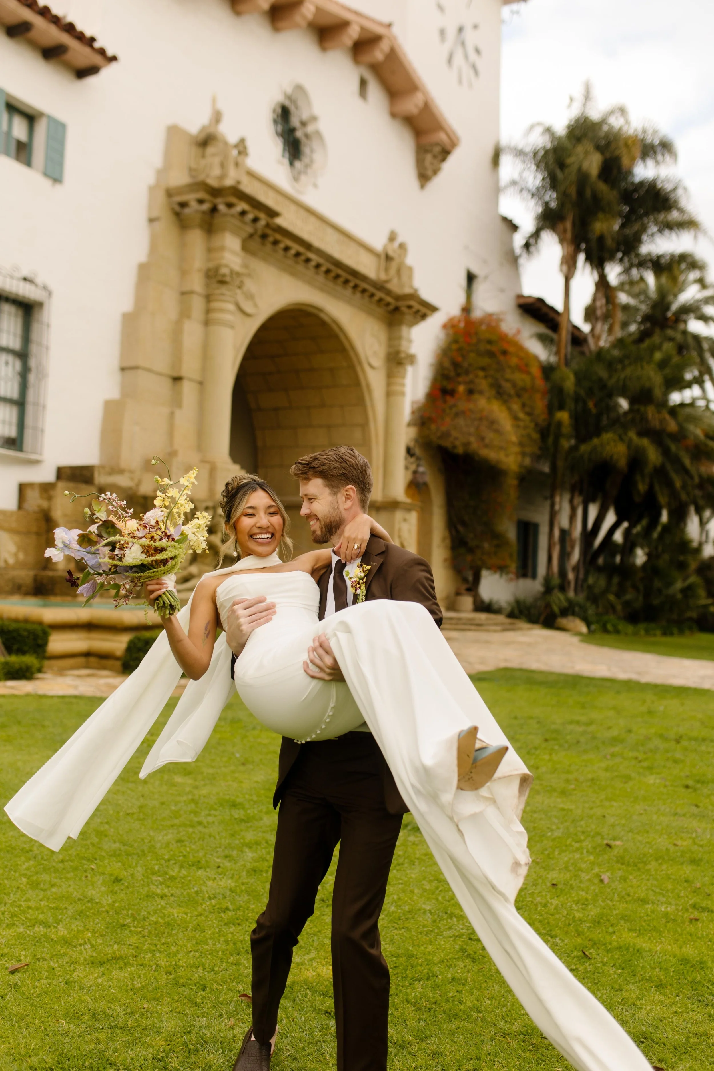 The groom lifts the bride off her feet on the lawn in front of the historic Santa Barbara Courthouse, both of them laughing in pure joy.