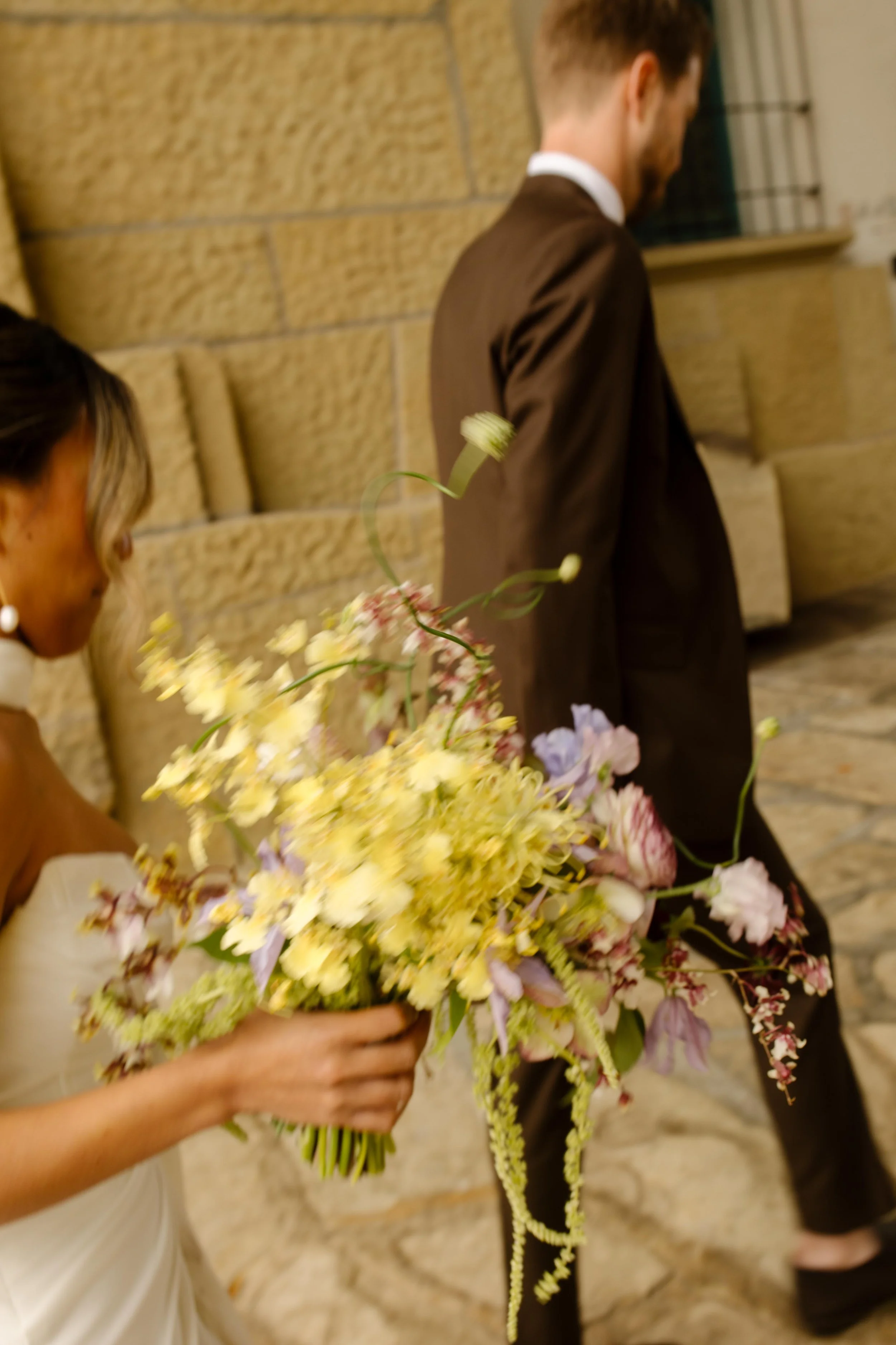The bride walks just behind the groom, bouquet in hand and veil catching in the breeze.