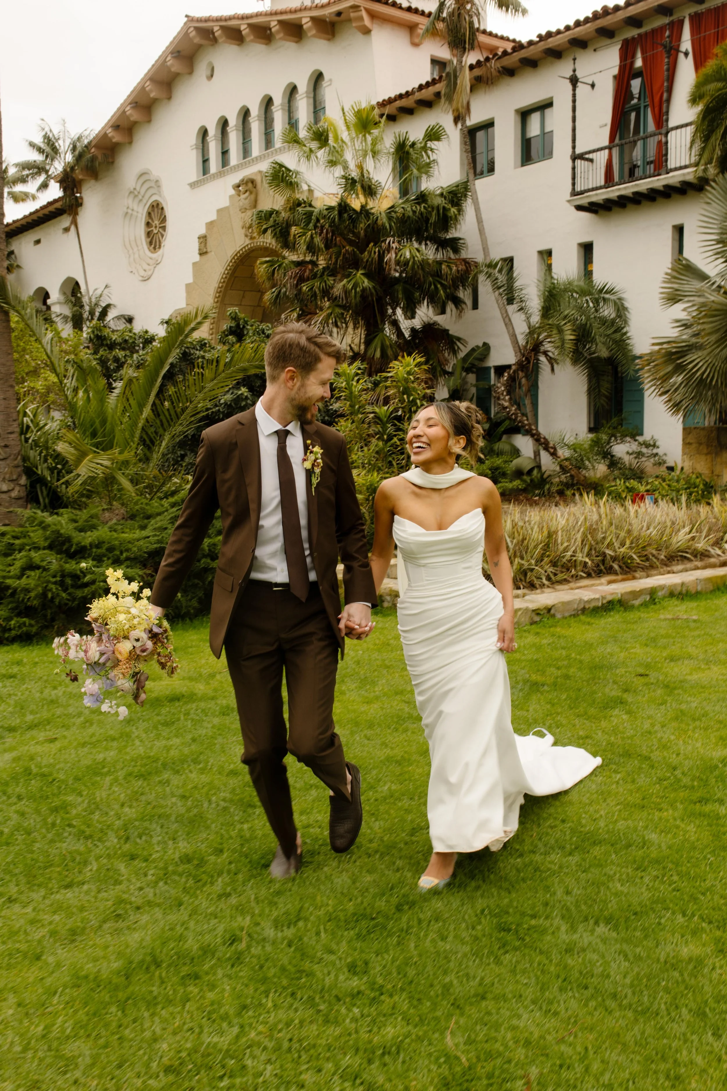 The couple walks hand in hand across the lawn, laughing in front of the iconic arches at their wedding Santa Barbara Courthouse.