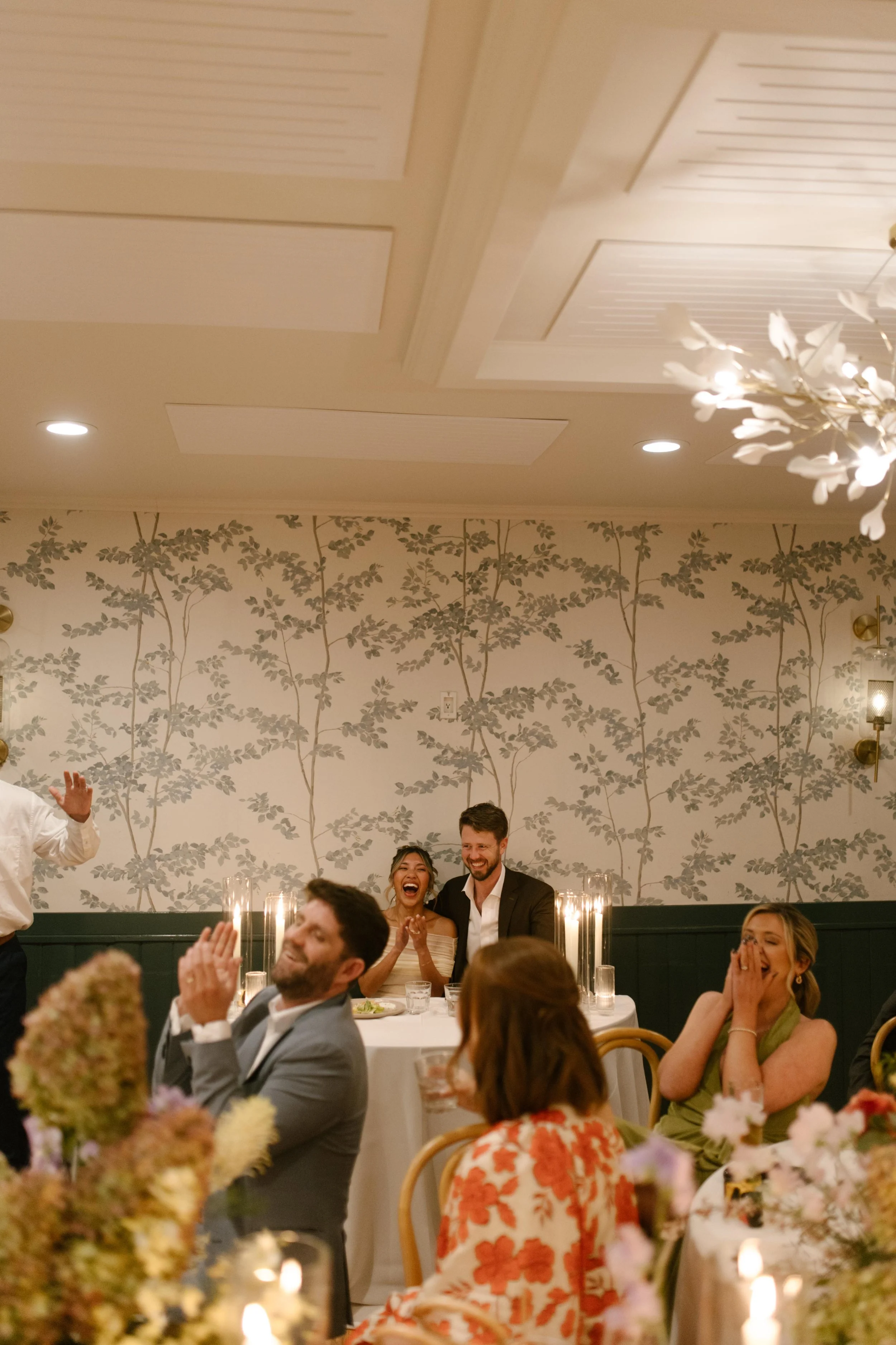 The couple sits at their sweetheart table, laughing as loved ones cheer during toasts.