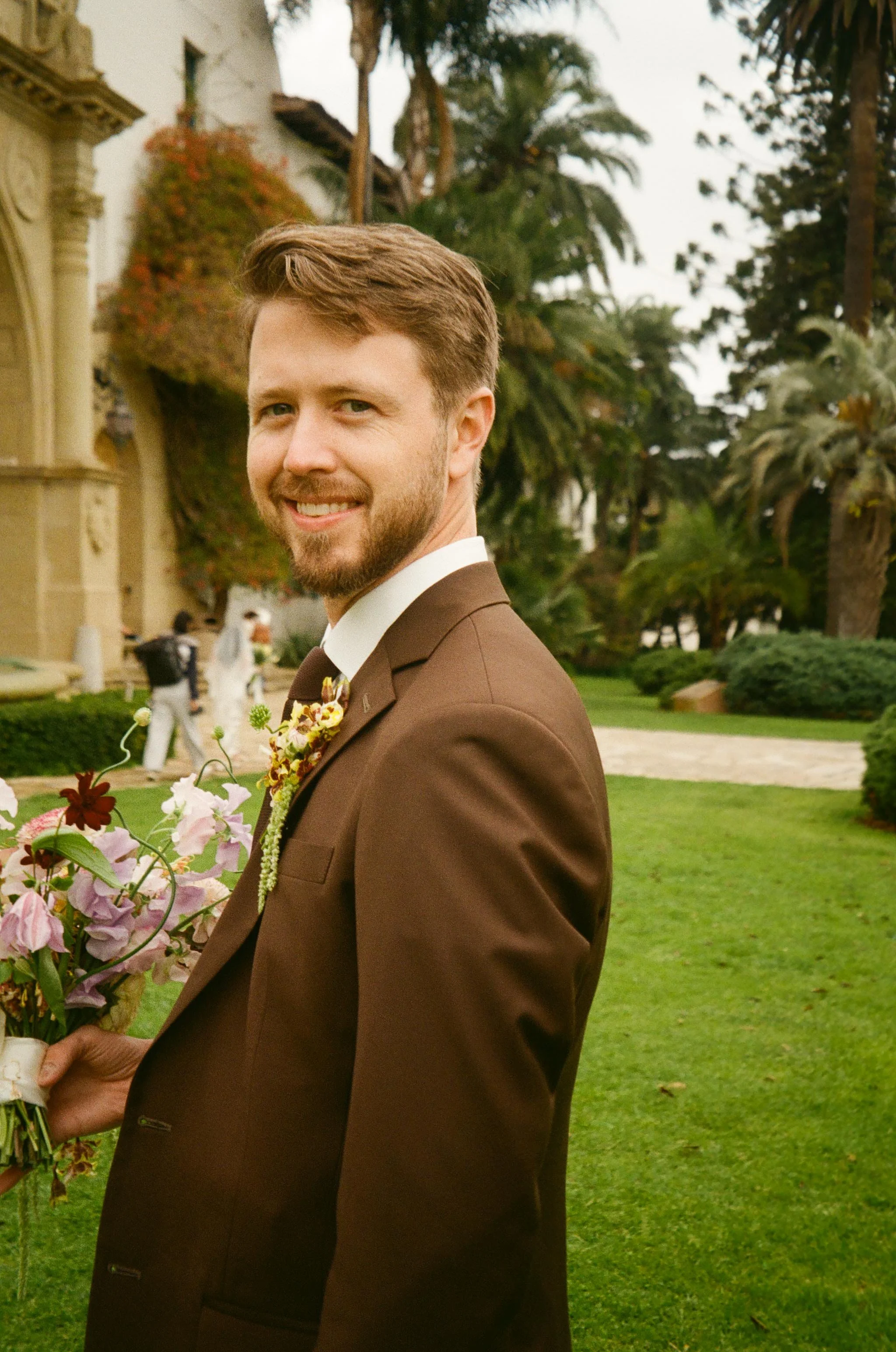 The groom smiles while holding the bouquet on courthouse grounds.