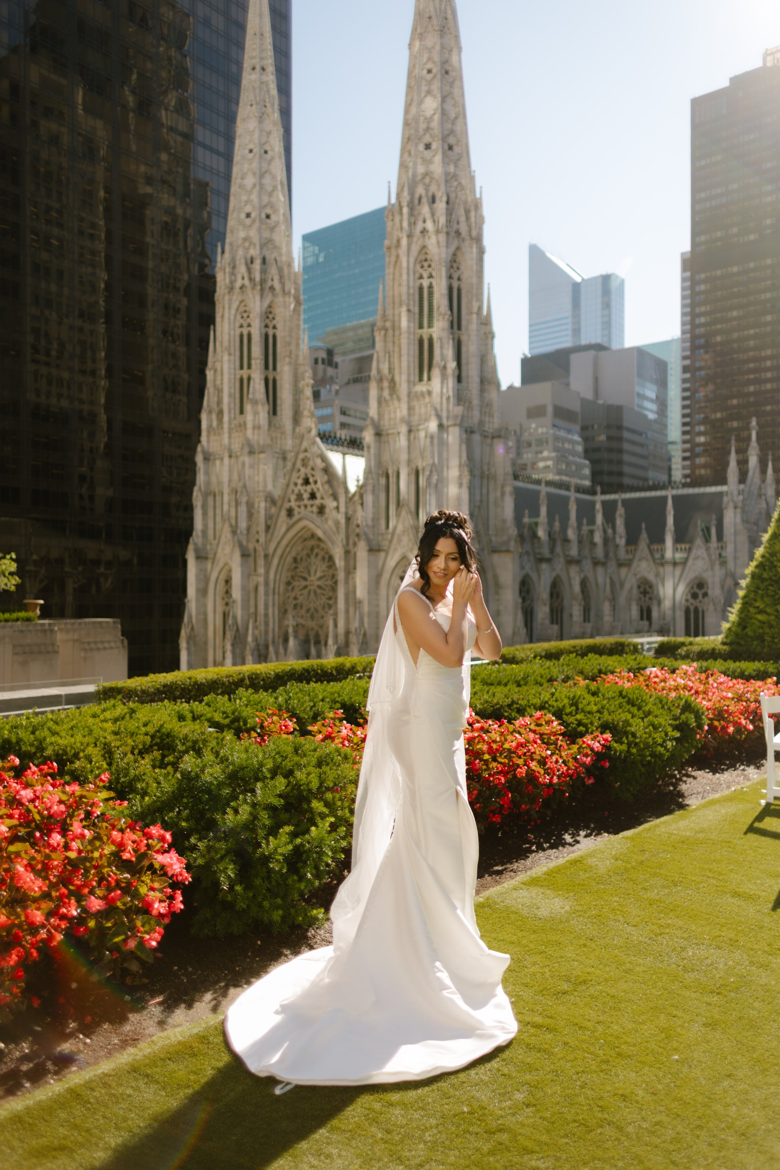 Bride standing in the sun-drenched rooftop garden, putting on her earrings with a soft smile—St. Patrick’s Cathedral rising behind her as she prepares for her wedding in New York City.