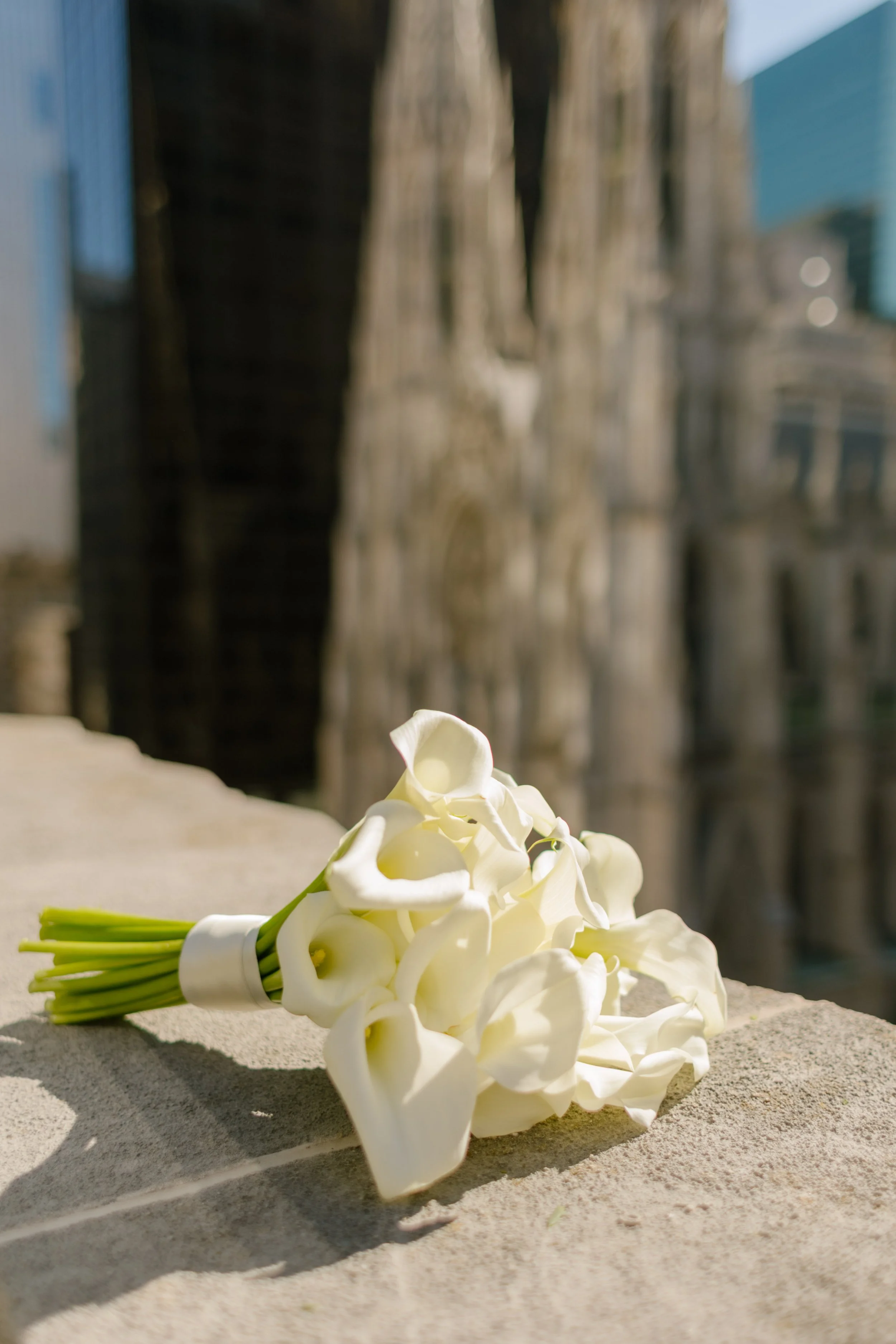 A simple and elegant bouquet of white calla lilies resting on a stone ledge, with blurred cathedral spires in the distance—an iconic detail from a wedding in New York City.