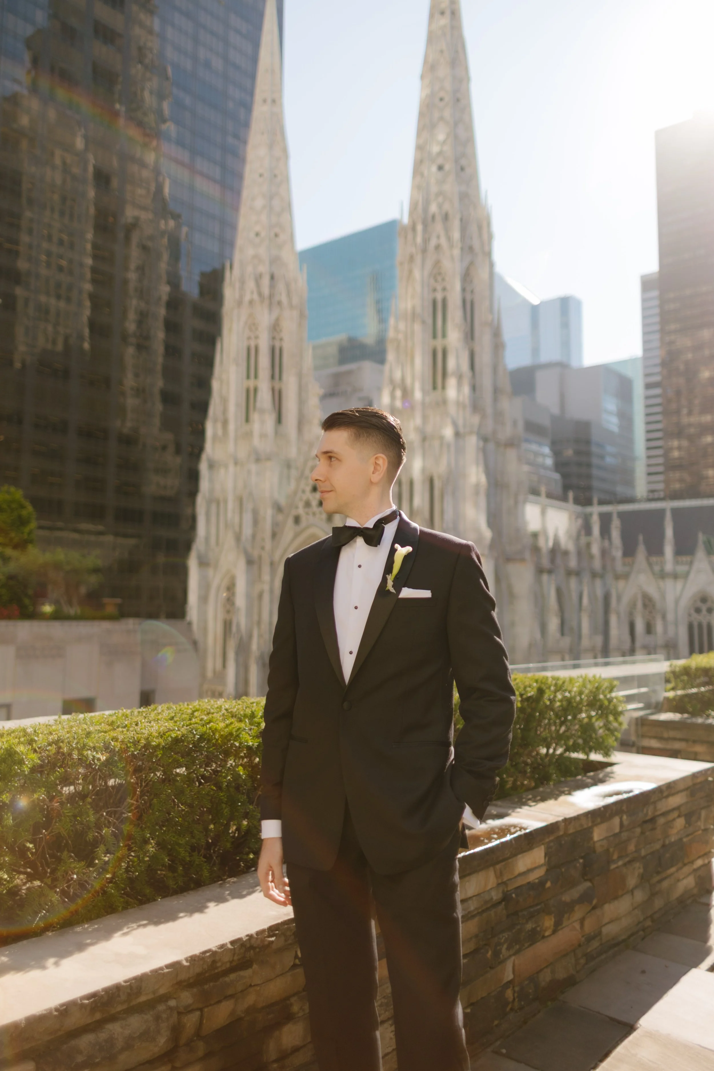 Groom in a classic black tuxedo standing on a rooftop garden, St. Patrick’s Cathedral glowing behind him in the morning light—a timeless portrait from a wedding in New York City.