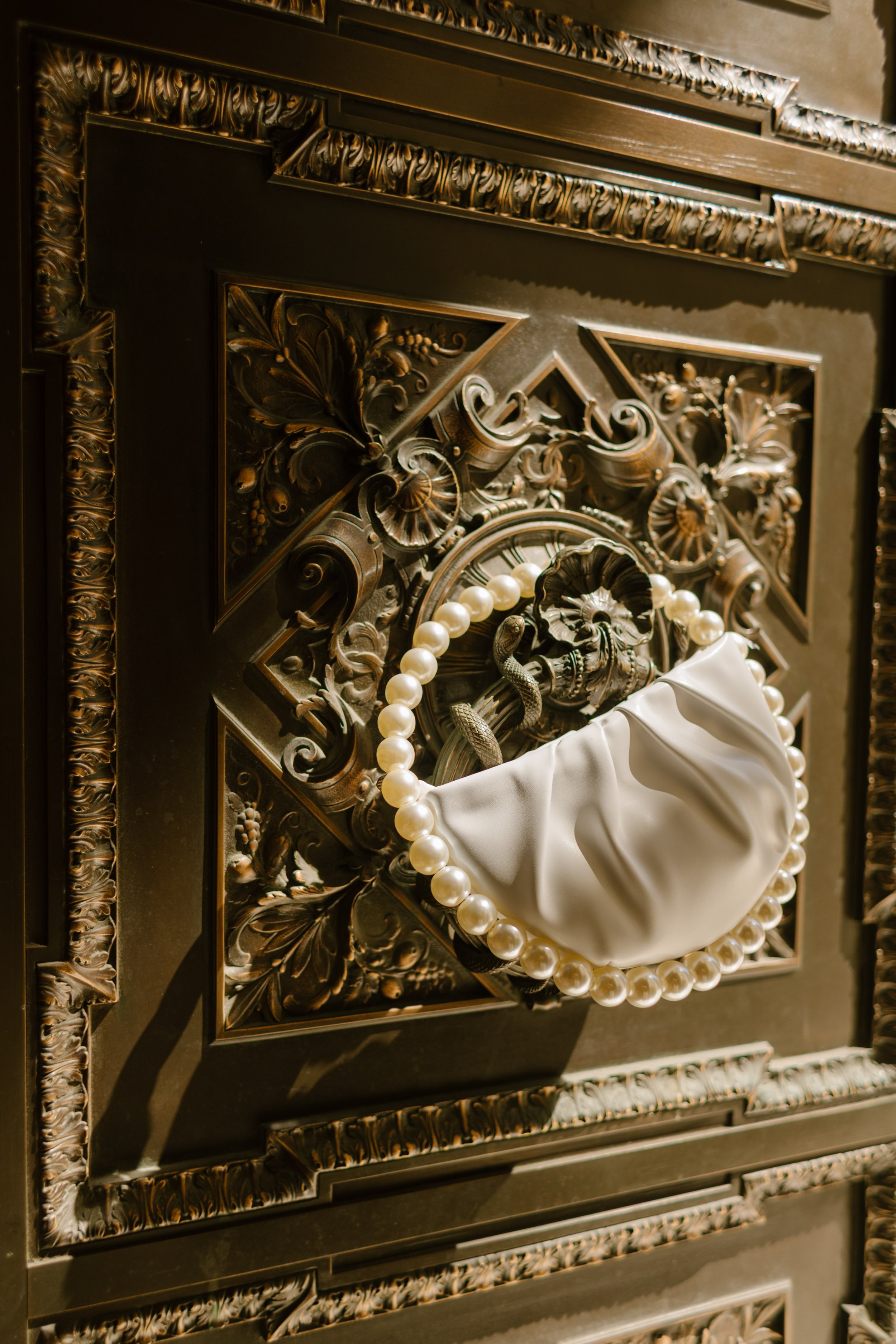 A pearl-handled bridal bag hanging on a vintage bronze door, catching golden afternoon light—a quiet detail full of old New York charm before a wedding in New York City.