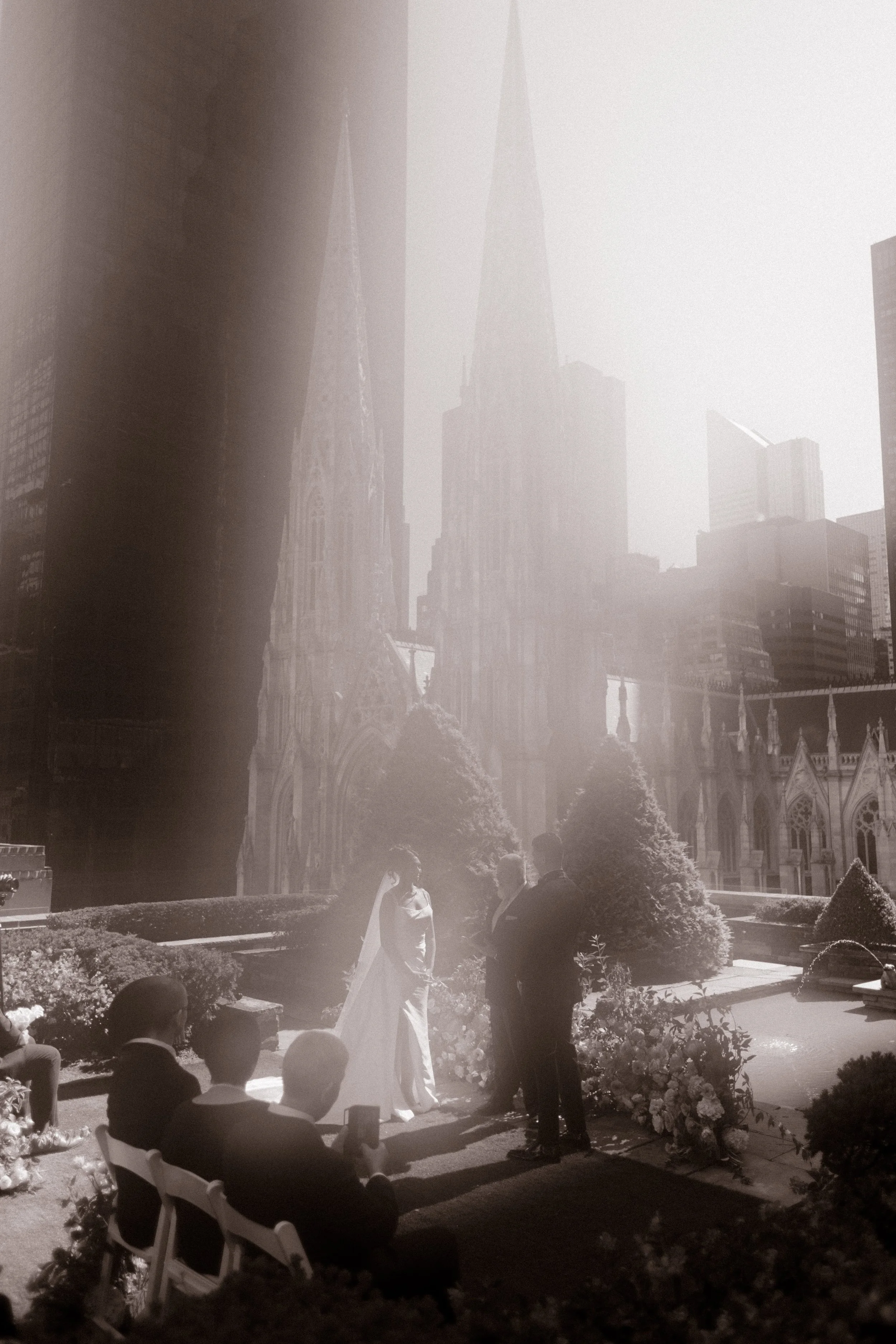 Soft, ethereal black-and-white image of the bride and groom exchanging vows on a rooftop garden, city skyline and cathedral rising behind the haze—a quiet pause in their wedding in New York City.