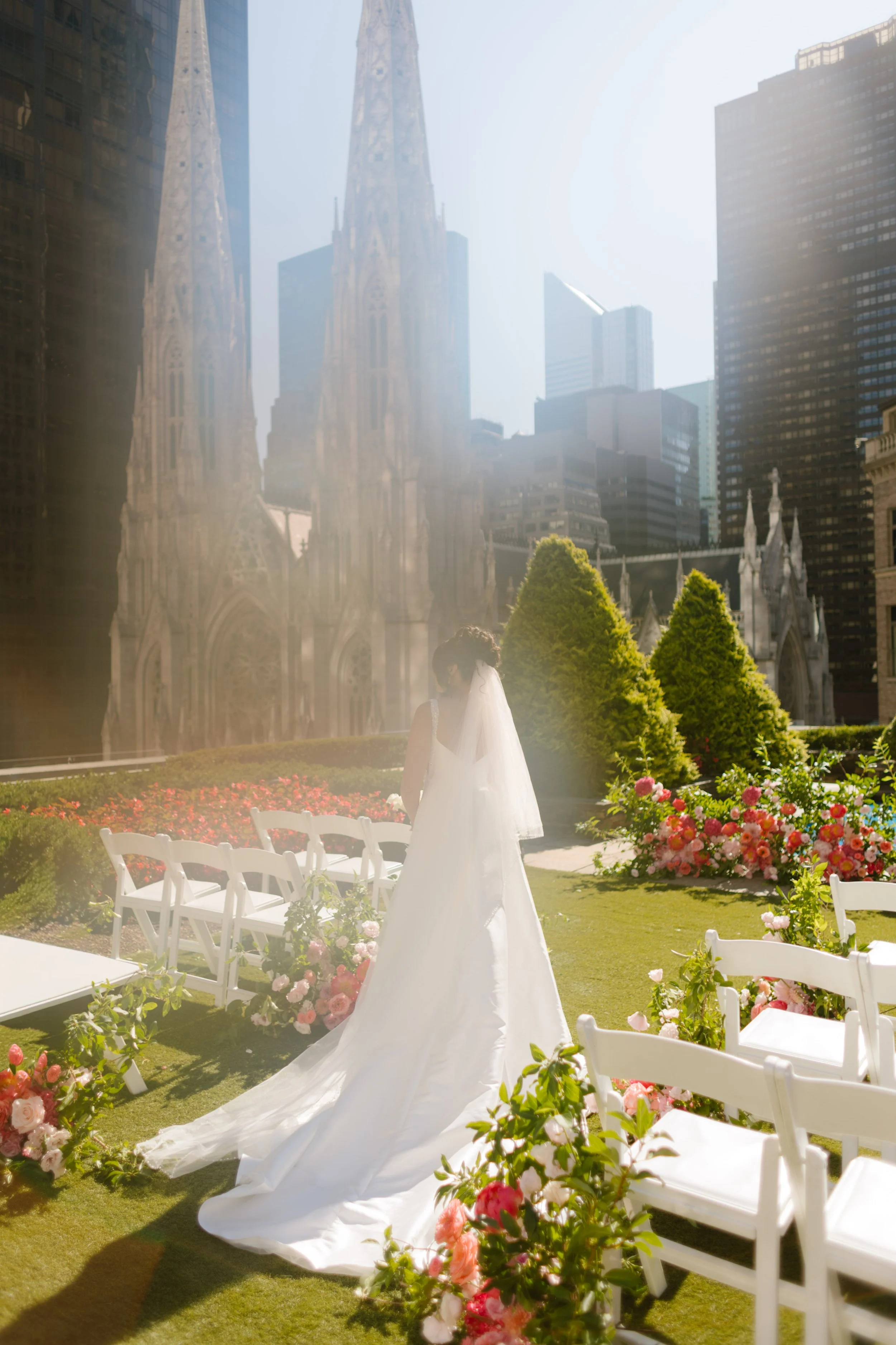 The bride walks alone through a golden haze toward the ceremony space, long veil and train trailing behind her as skyscrapers glow around—an iconic entrance to her wedding in New York City.