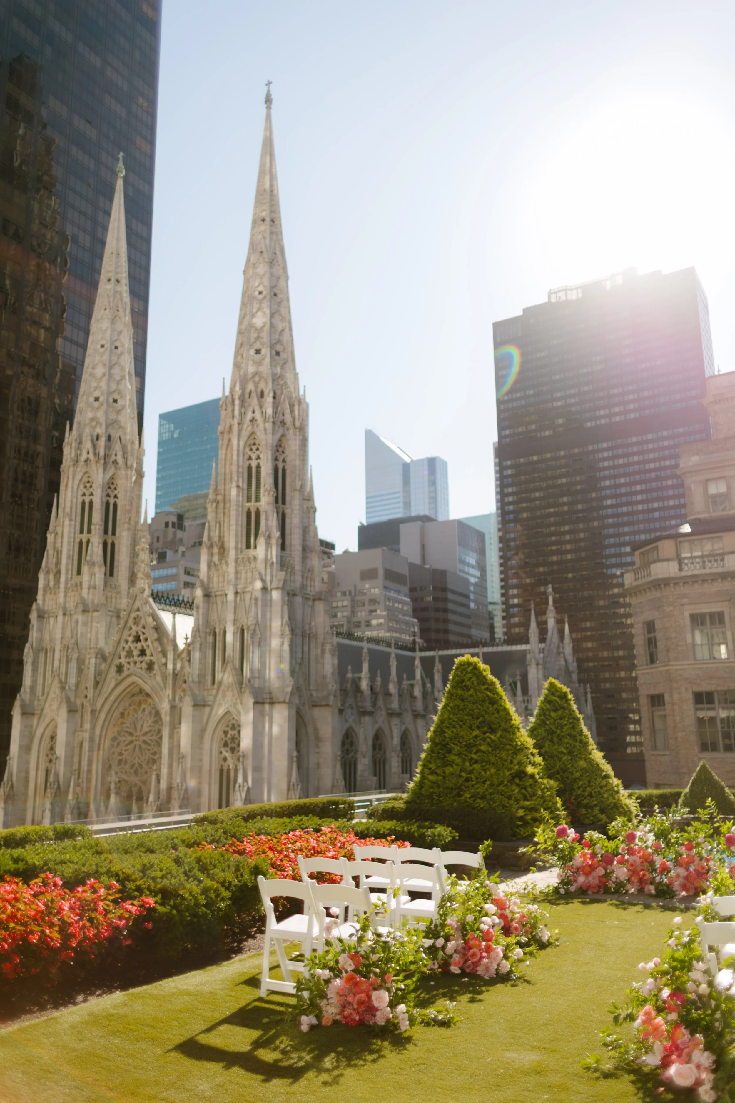 White ceremony chairs lined with lush pink florals on a rooftop garden, with St. Patrick’s Cathedral and NYC skyscrapers rising in the background—a dreamy setup for a wedding in New York City.