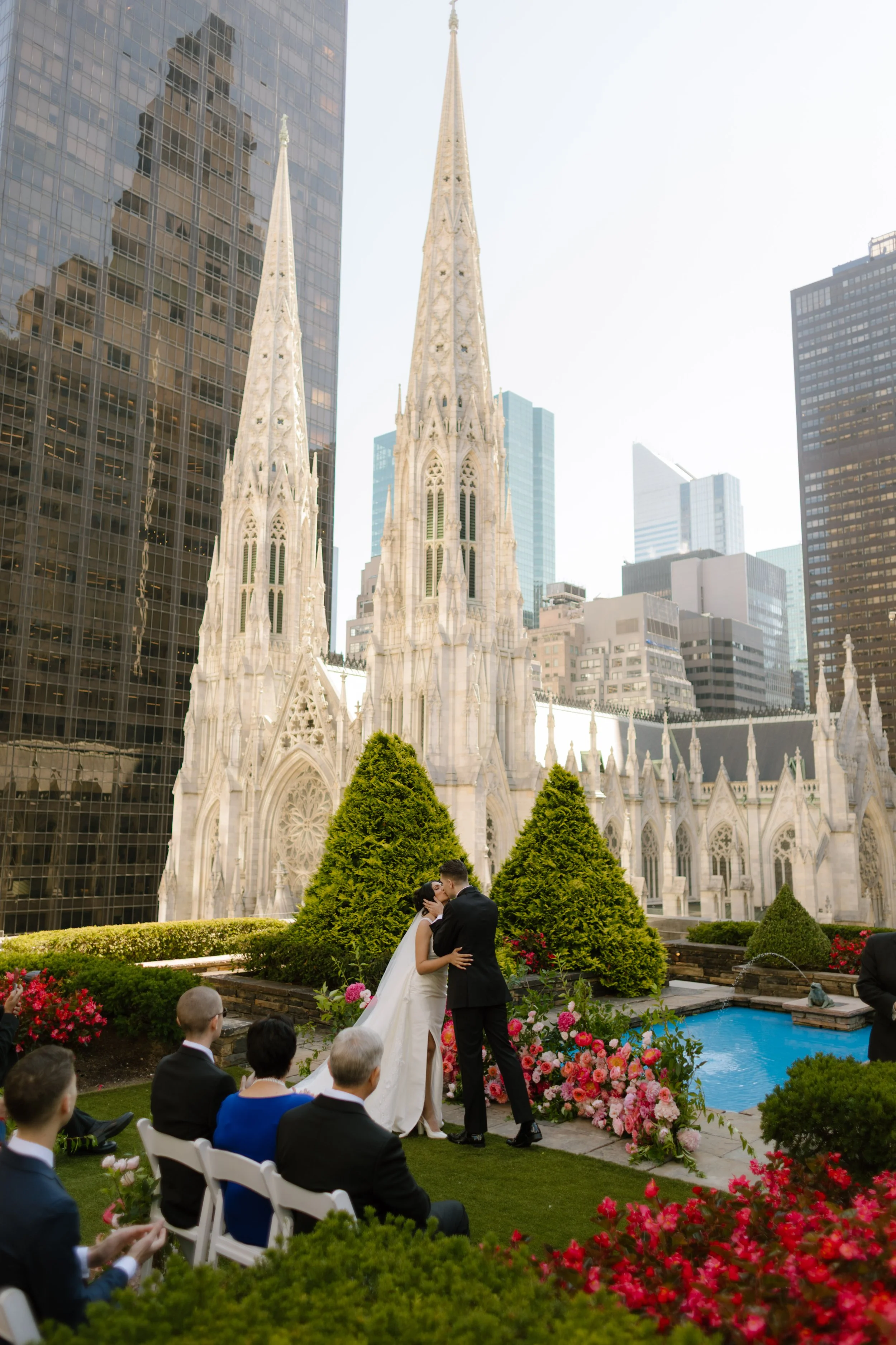 he bride and groom share their first kiss in front of guests on a rooftop garden bursting with florals, with St. Patrick’s Cathedral behind them