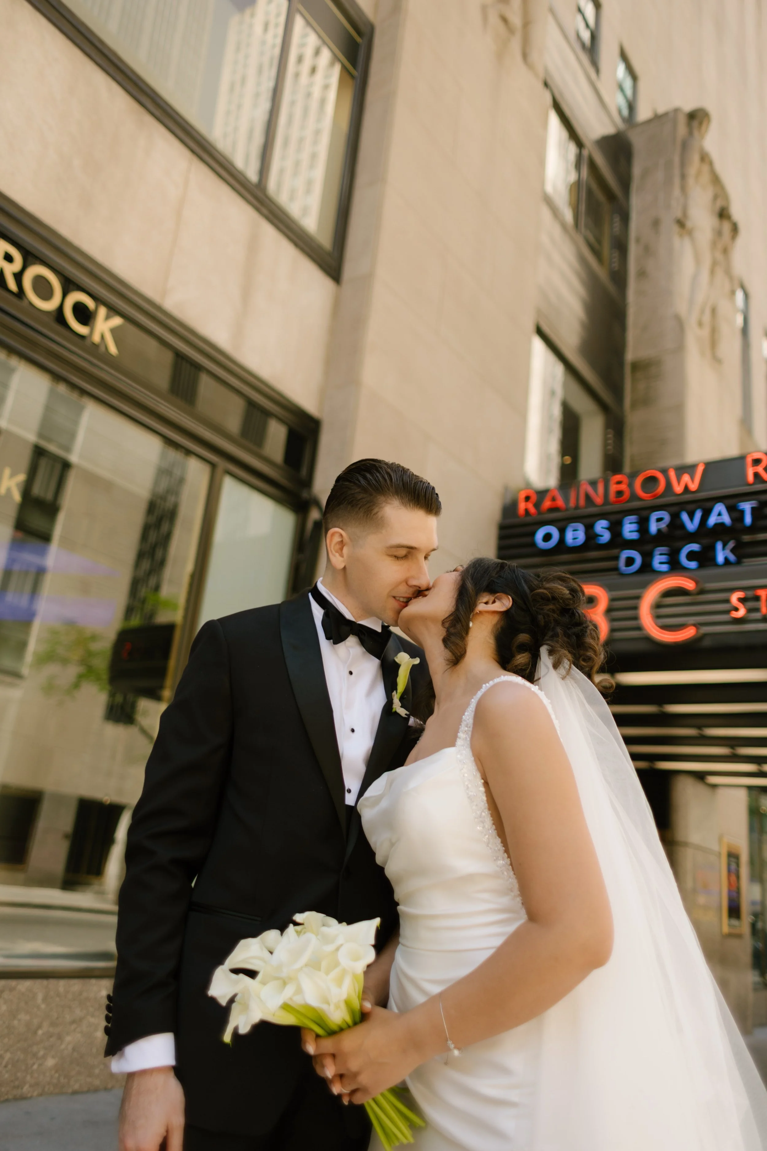 The bride kisses the groom’s cheek as they stand under the neon glow of the Rainbow Room sign
