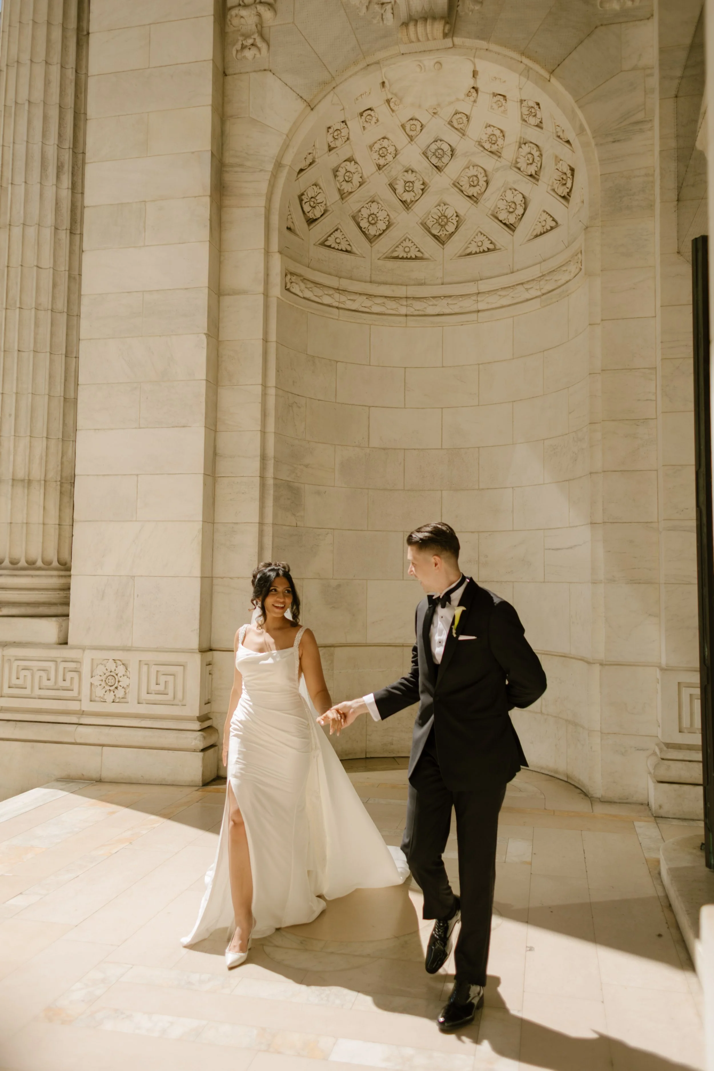 The bride and groom walk hand in hand through the grand marble halls of the New York Public Library