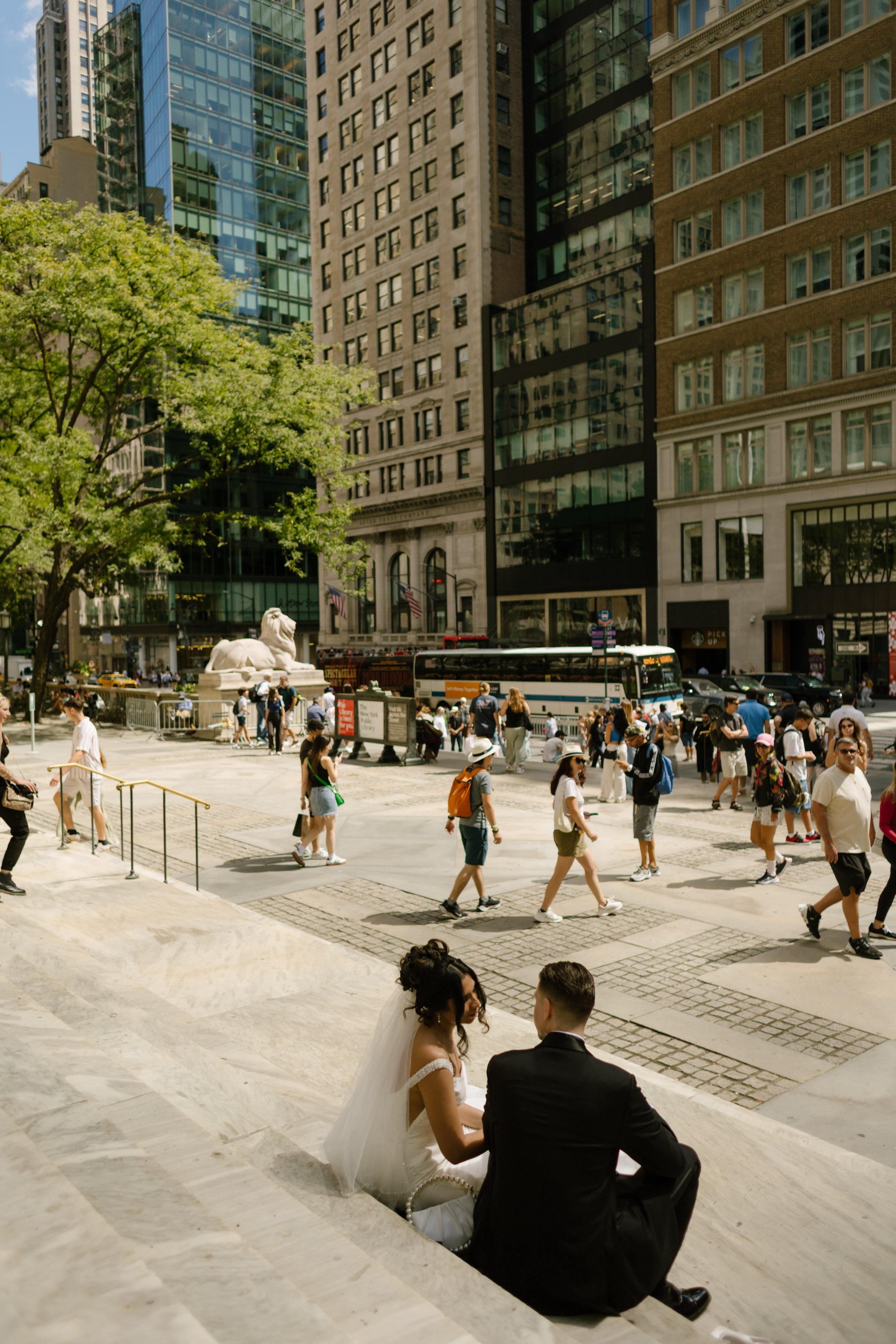 The bride and groom sit on the marble steps of the New York Public Library, tucked into a quiet moment together while the city moves around them—a soft pause in their wedding in New York City.