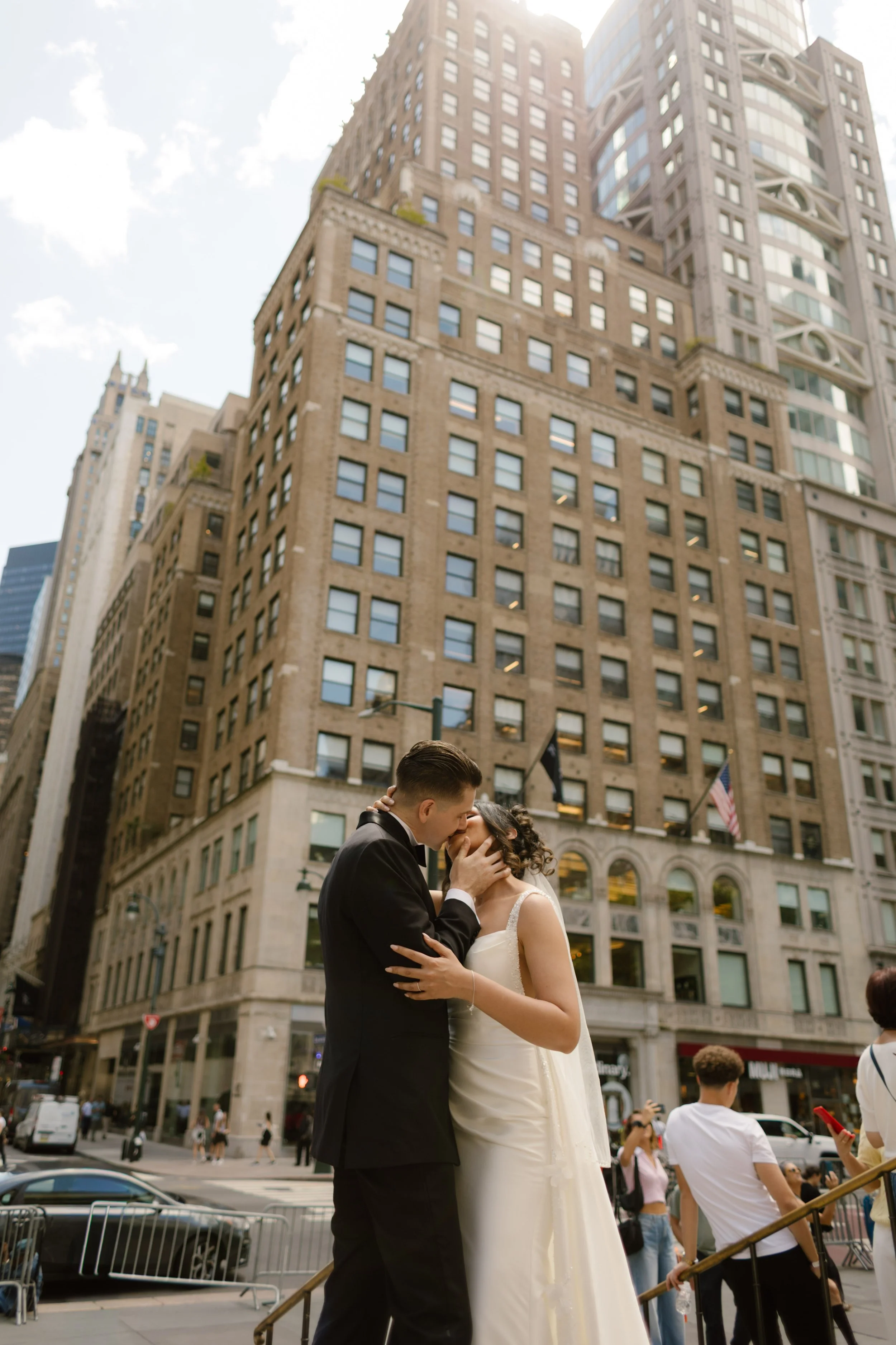 The groom cradles the bride’s face as they kiss on a bustling Midtown corner, skyscrapers glowing behind them