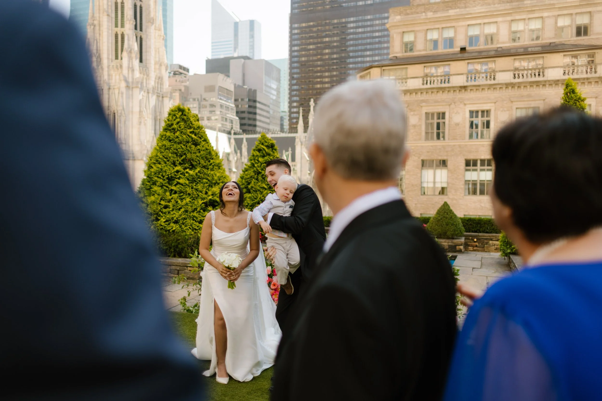 The bride throws her head back laughing while the groom holds their sleepy child, framed by family and the NYC skyline—a moment of real life and deep love during their wedding in New York City.