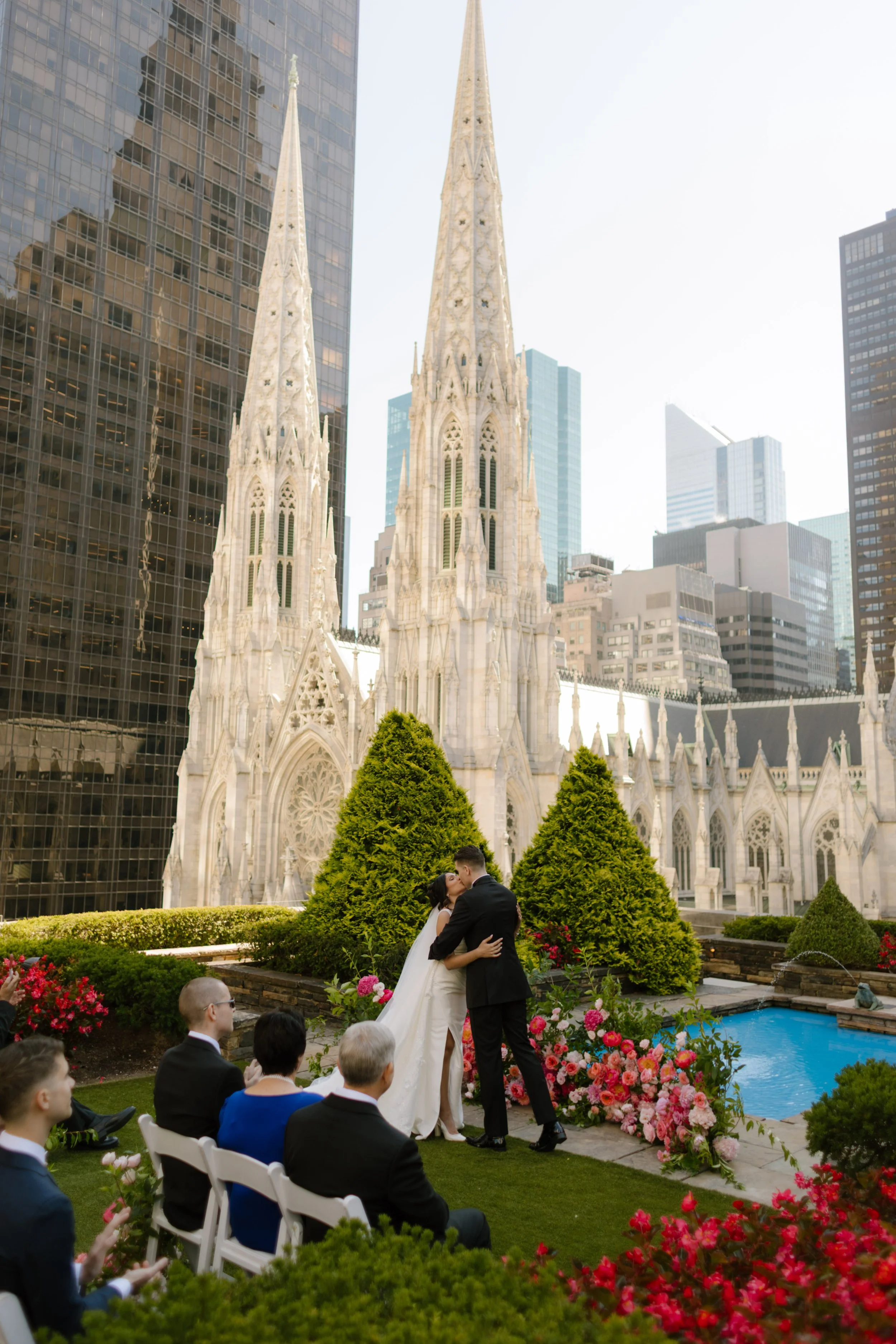 The newlyweds share their first kiss as husband and wife, surrounded by vibrant florals and loved ones on a rooftop overlooking St. Patrick’s—pure magic during their wedding in New York City.
