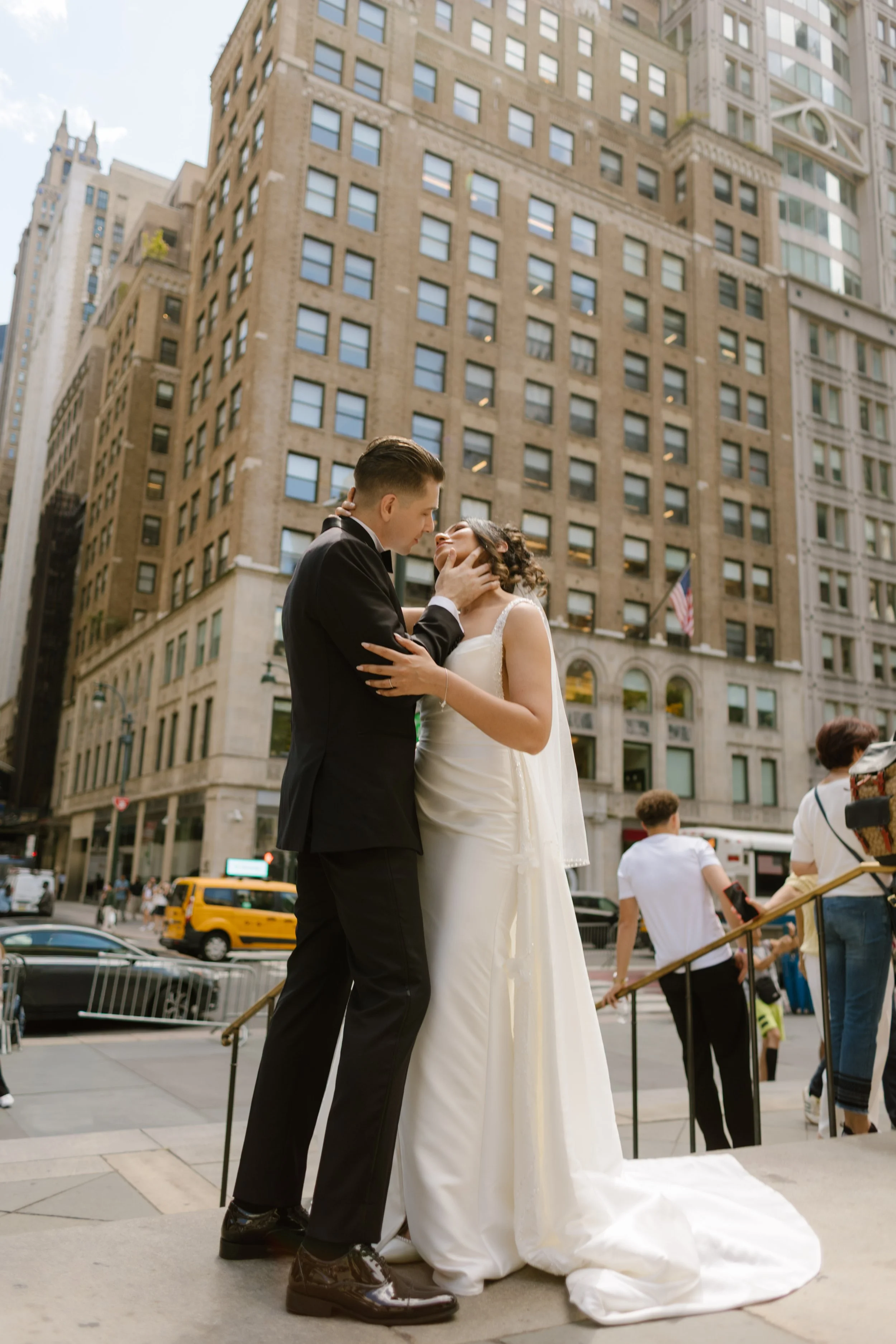 A playful, romantic moment as the groom holds the bride’s face in both hands on the city steps—surrounded by yellow cabs and strangers on the move,
