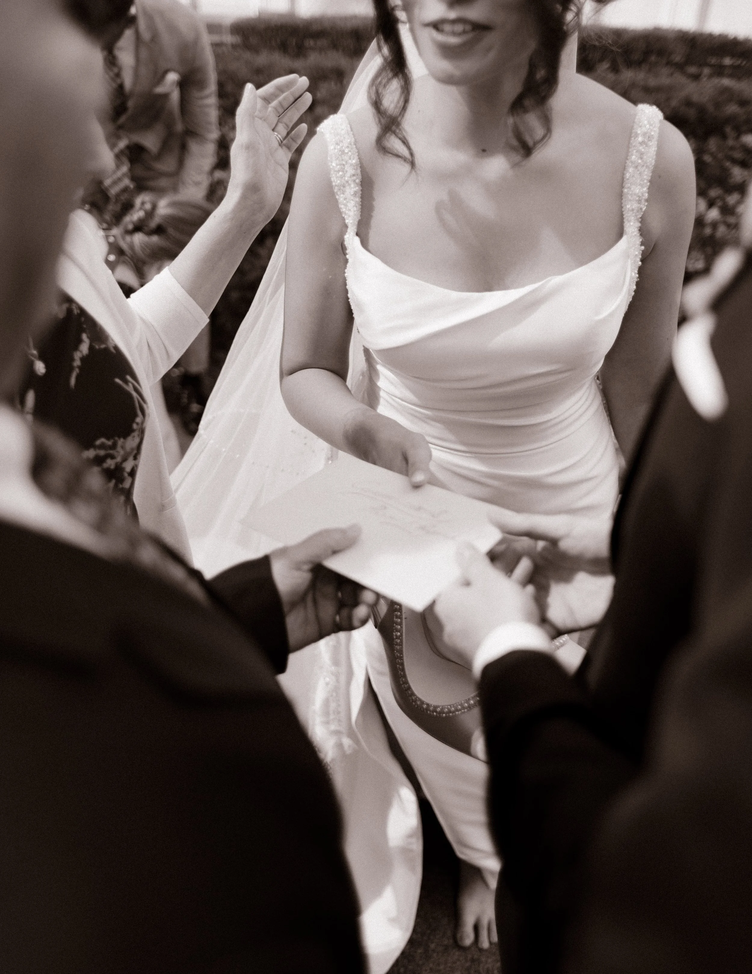 A sepia-toned candid of the bride accepting a letter, barefoot and smiling, surrounded by hands and love