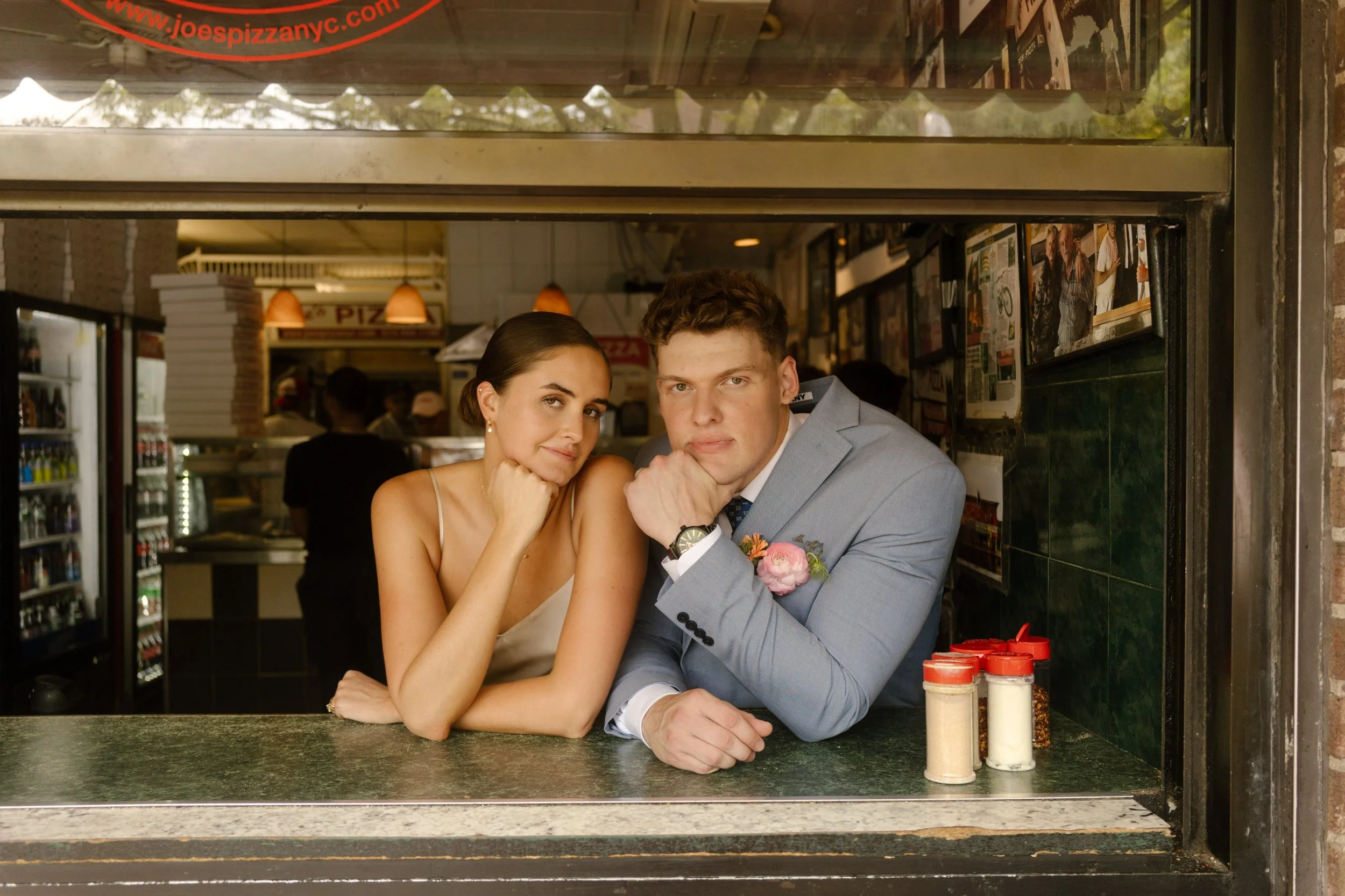 Bride and groom leaning through the window counter at Joe’s Pizza, sharing a playful, candid moment during their New York City elopement.