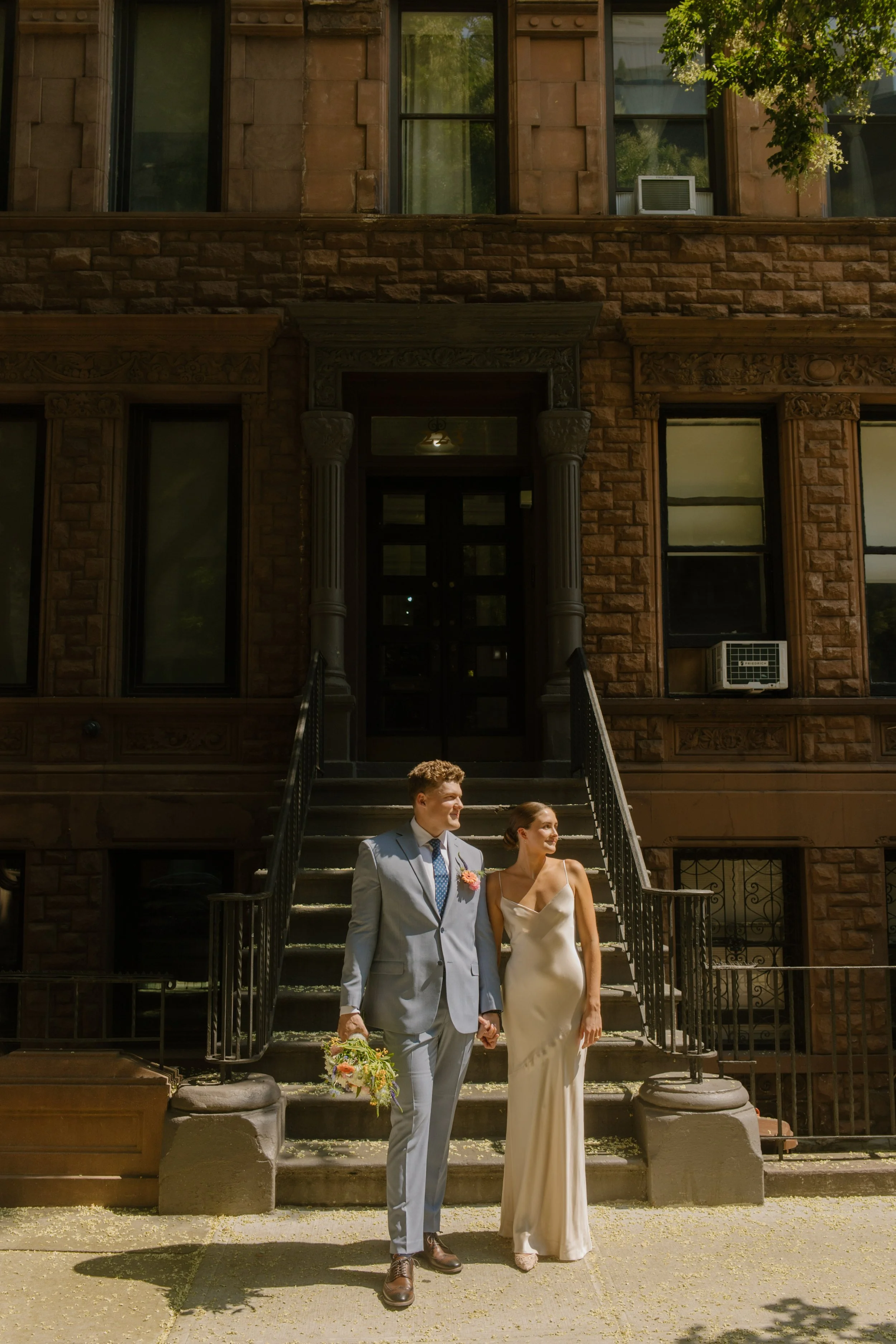 Bride and groom standing hand-in-hand at the base of a brownstone staircase, framed by soft shadows and sunlit stone.