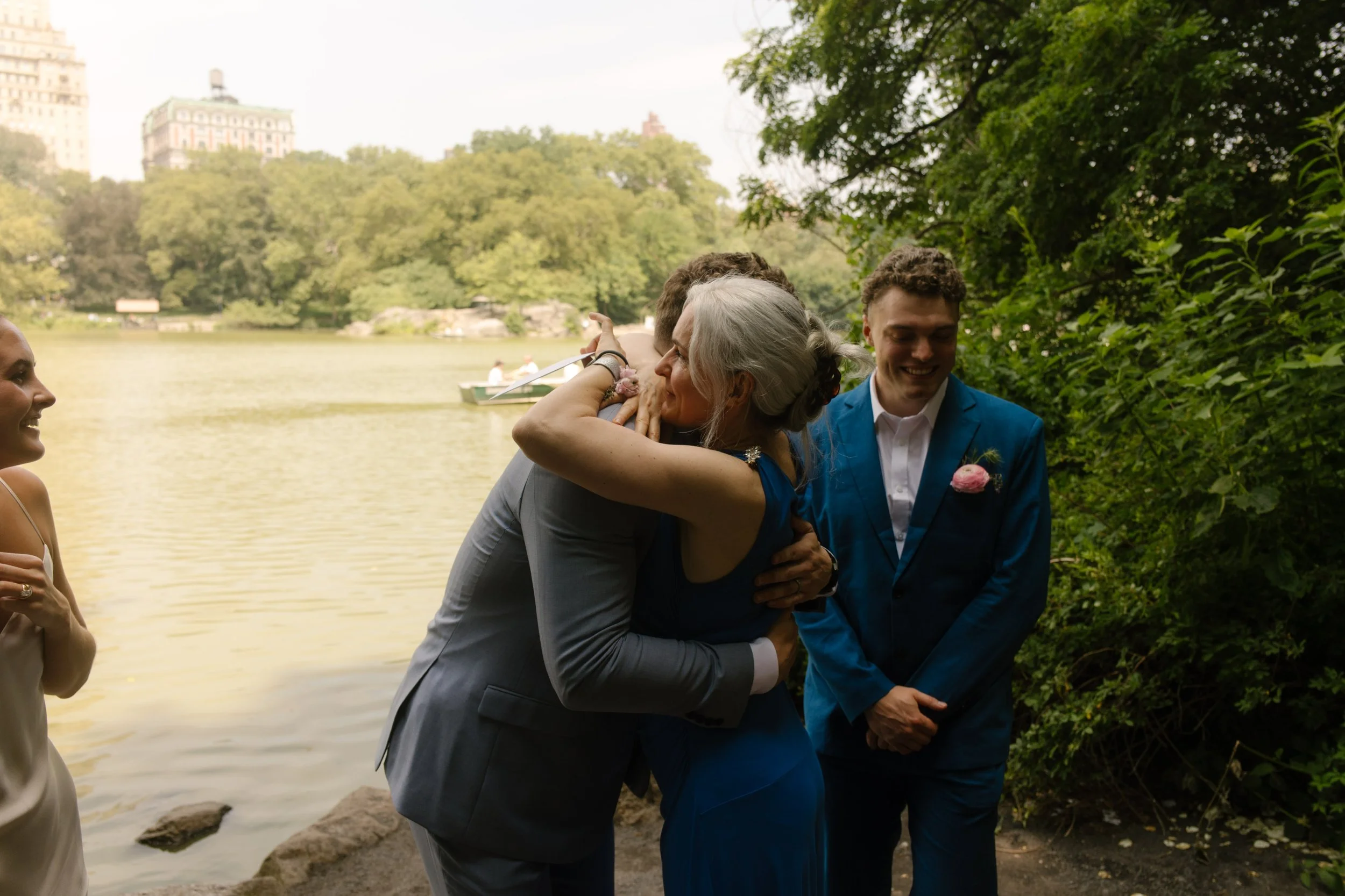 The groom embraces a family member near the water, as rowboats drift by behind them in this emotional post-ceremony moment.