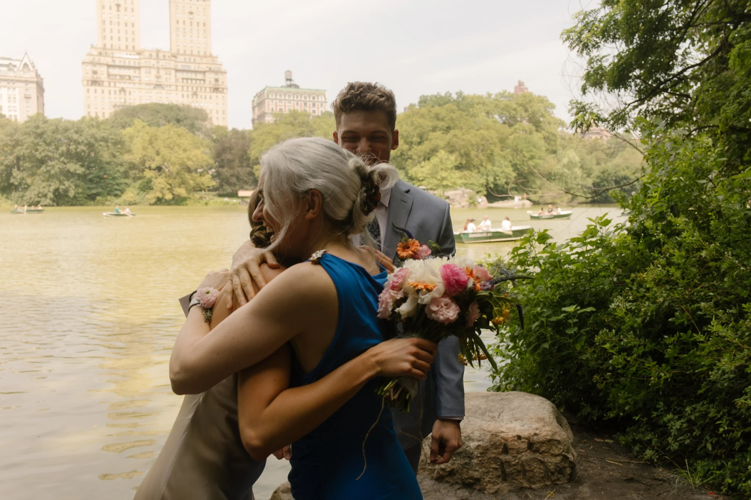 The bride hugs a loved one, bouquet in hand, with the lake and iconic Manhattan buildings glowing behind her during this New York City elopement celebration.