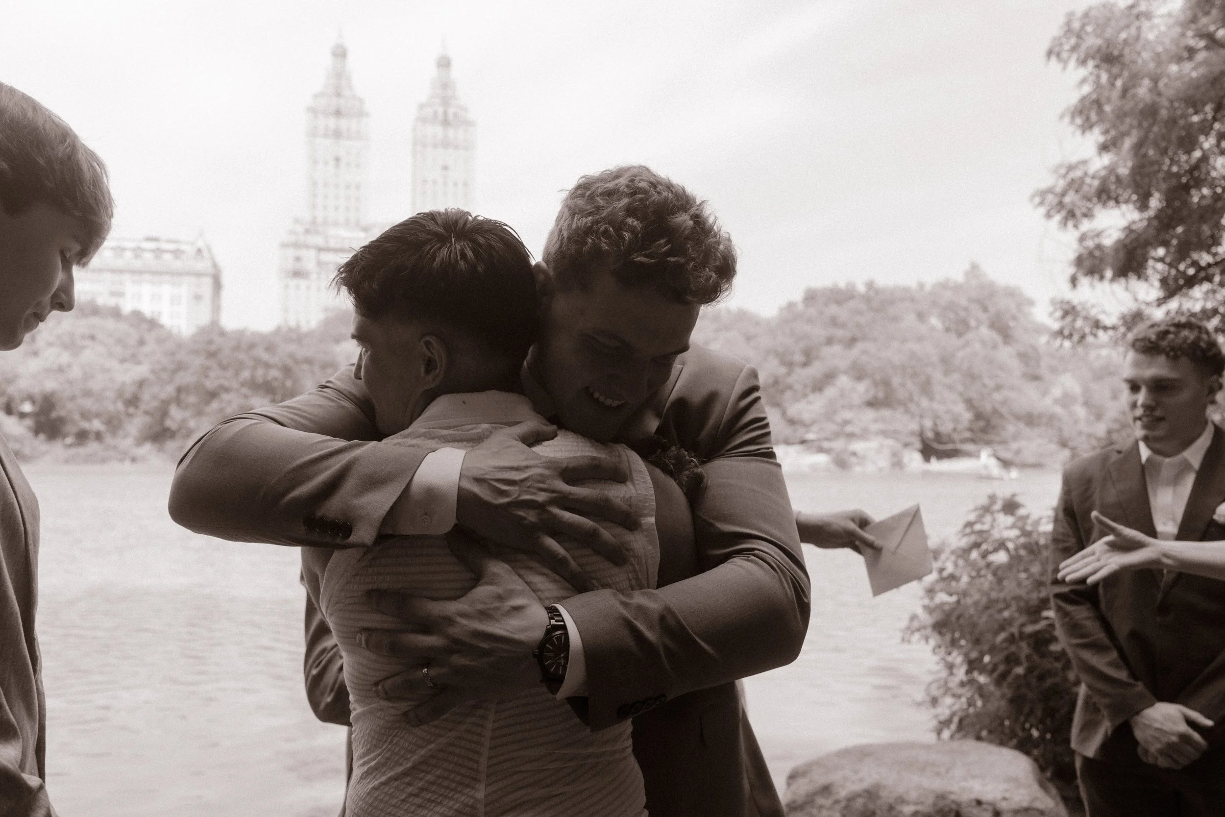 A tearful black and white hug between the groom and a friend, with the Central Park lake and NYC skyline in the background.