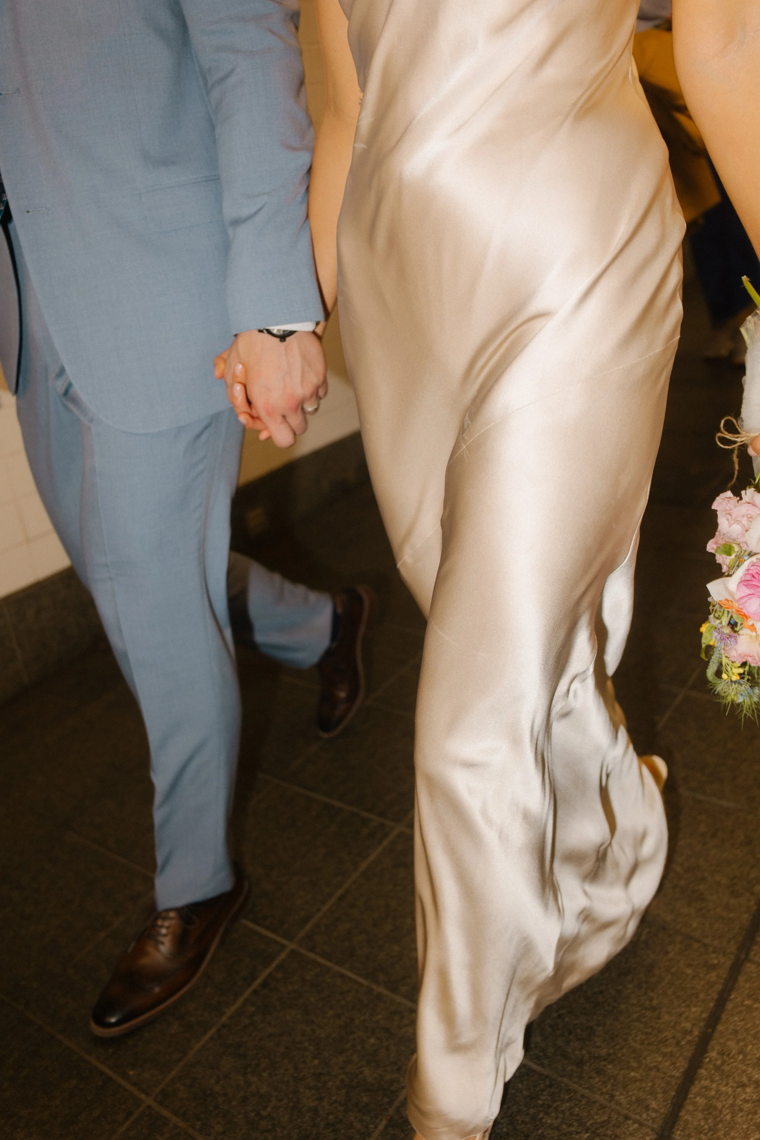 Close-up of the couple holding hands while walking through a subway station, silk dress swaying and bouquet in motion.