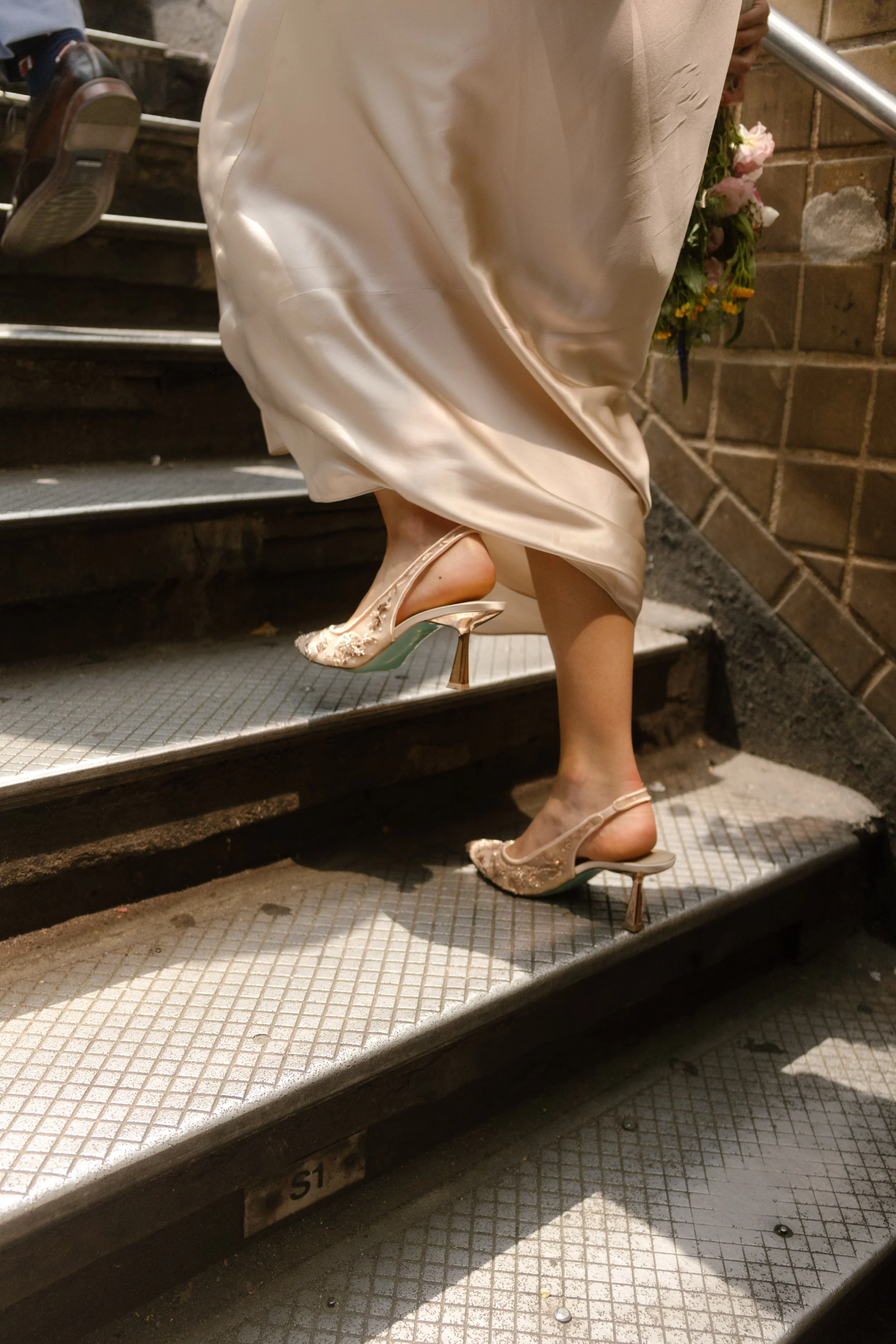Bride walking up subway stairs, dress flowing and heels glinting in the sun.