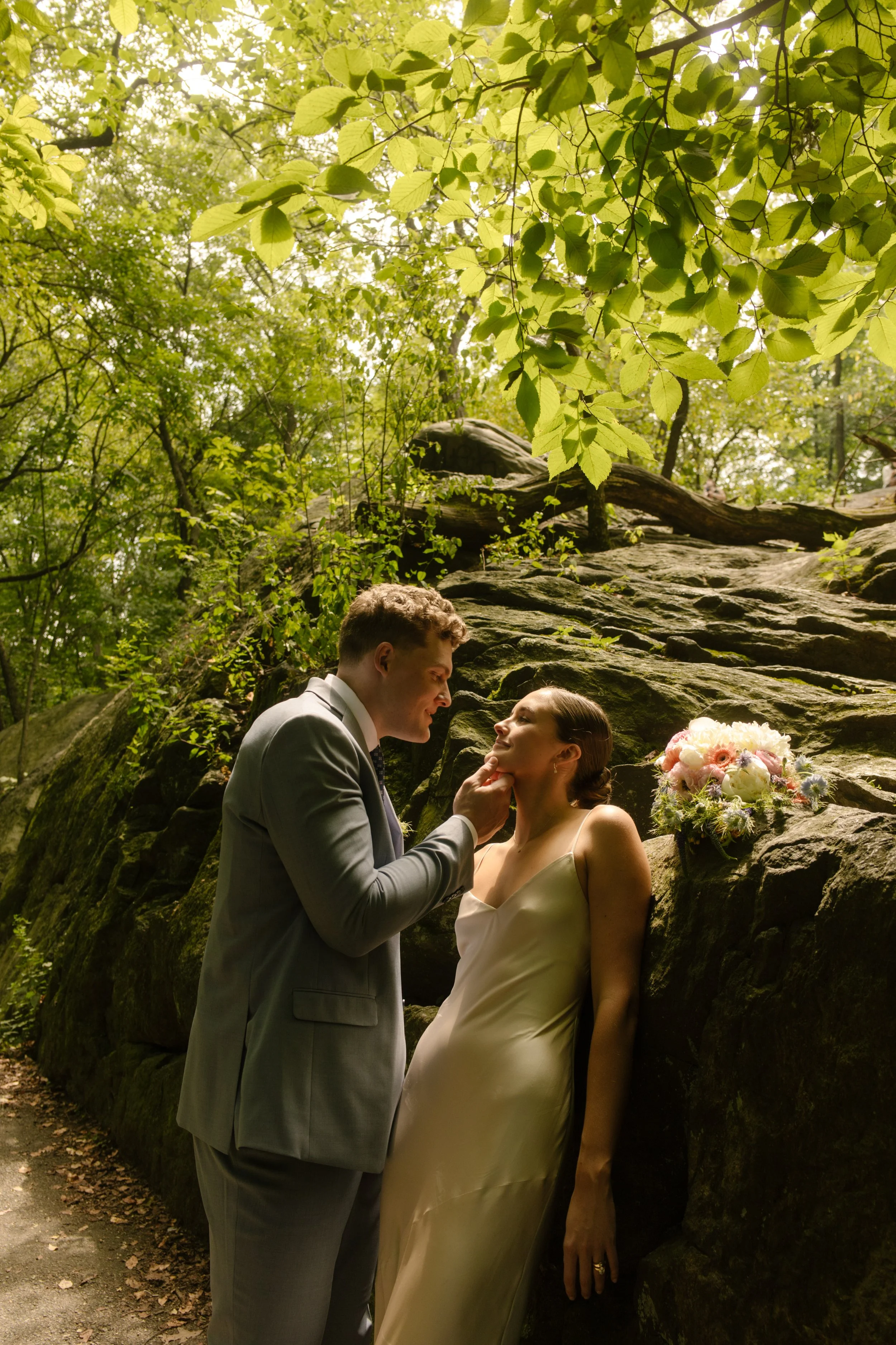 Tender moment in Central Park, with the groom holding the bride’s chin beside a bouquet resting on a rock.