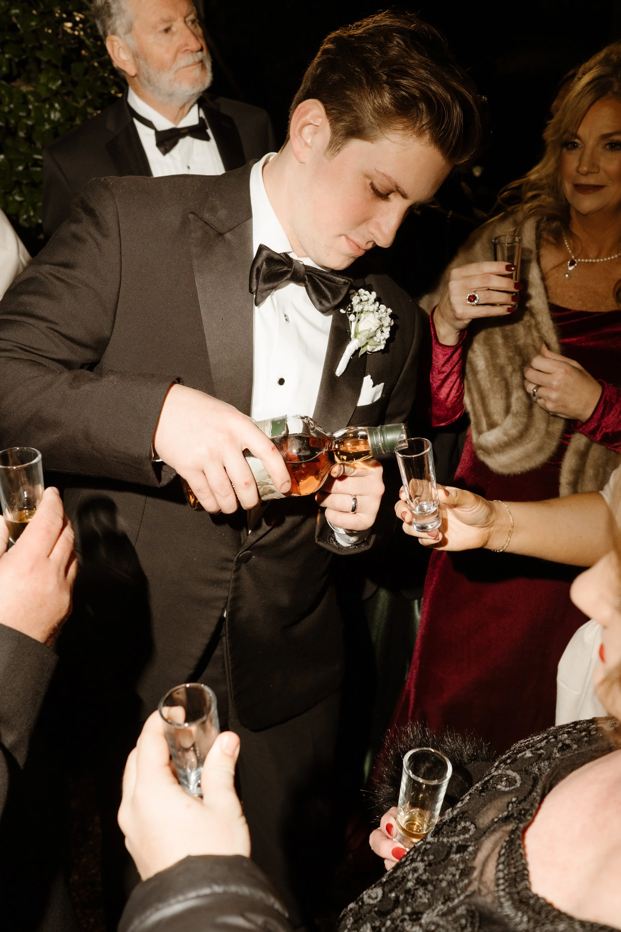Surrounded by guests, the groom pours shots with a focused hand and a festive crowd cheering him on.