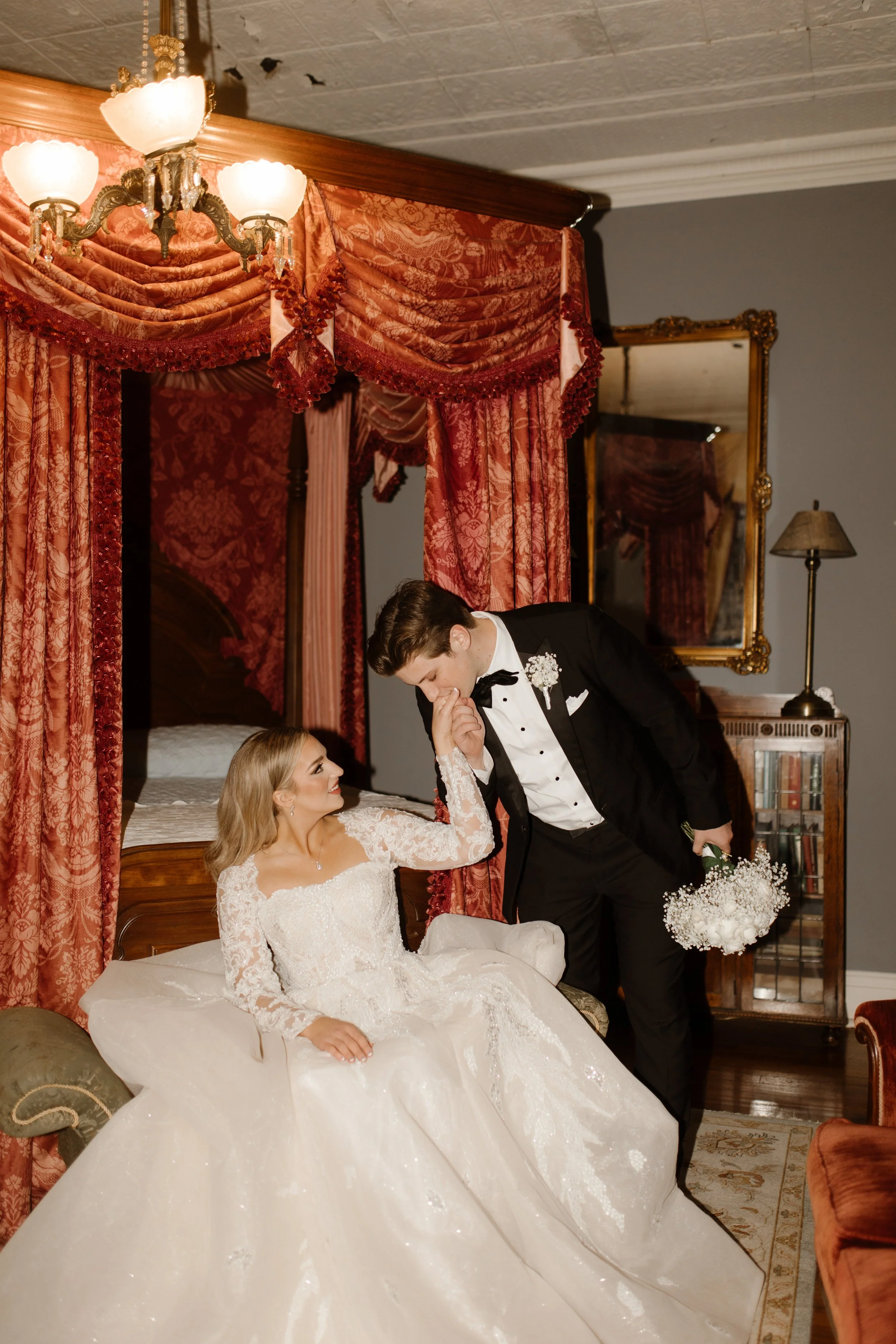 In a richly decorated suite, the groom leans down to kiss his bride’s hand while she sits in her gown, bouquet still in hand—pure old-Hollywood Arkansas wedding magic.