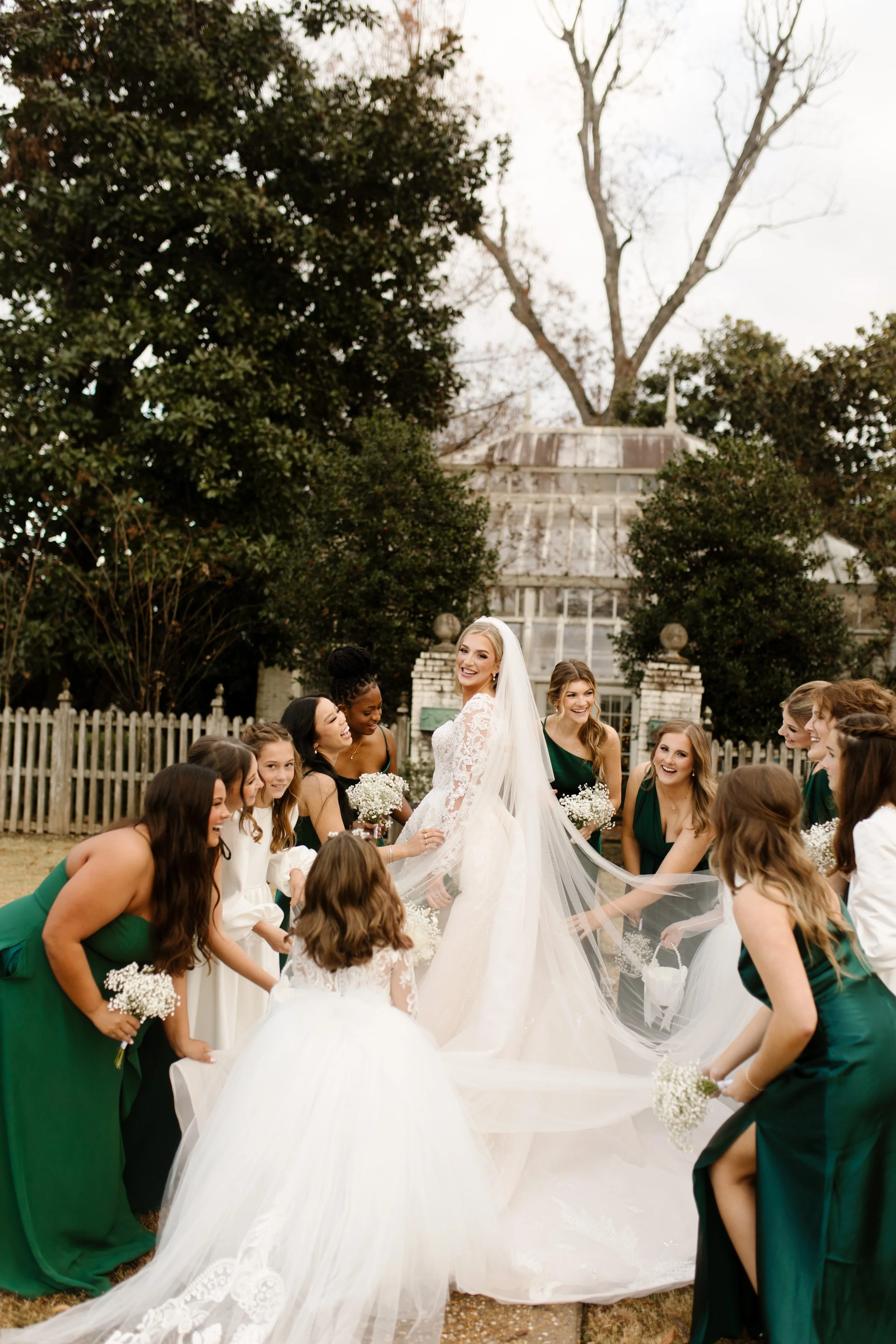 A joyful scene of bridesmaids gathered around the bride, fluffing her veil and train outside a greenhouse before the Arkansas wedding ceremony.