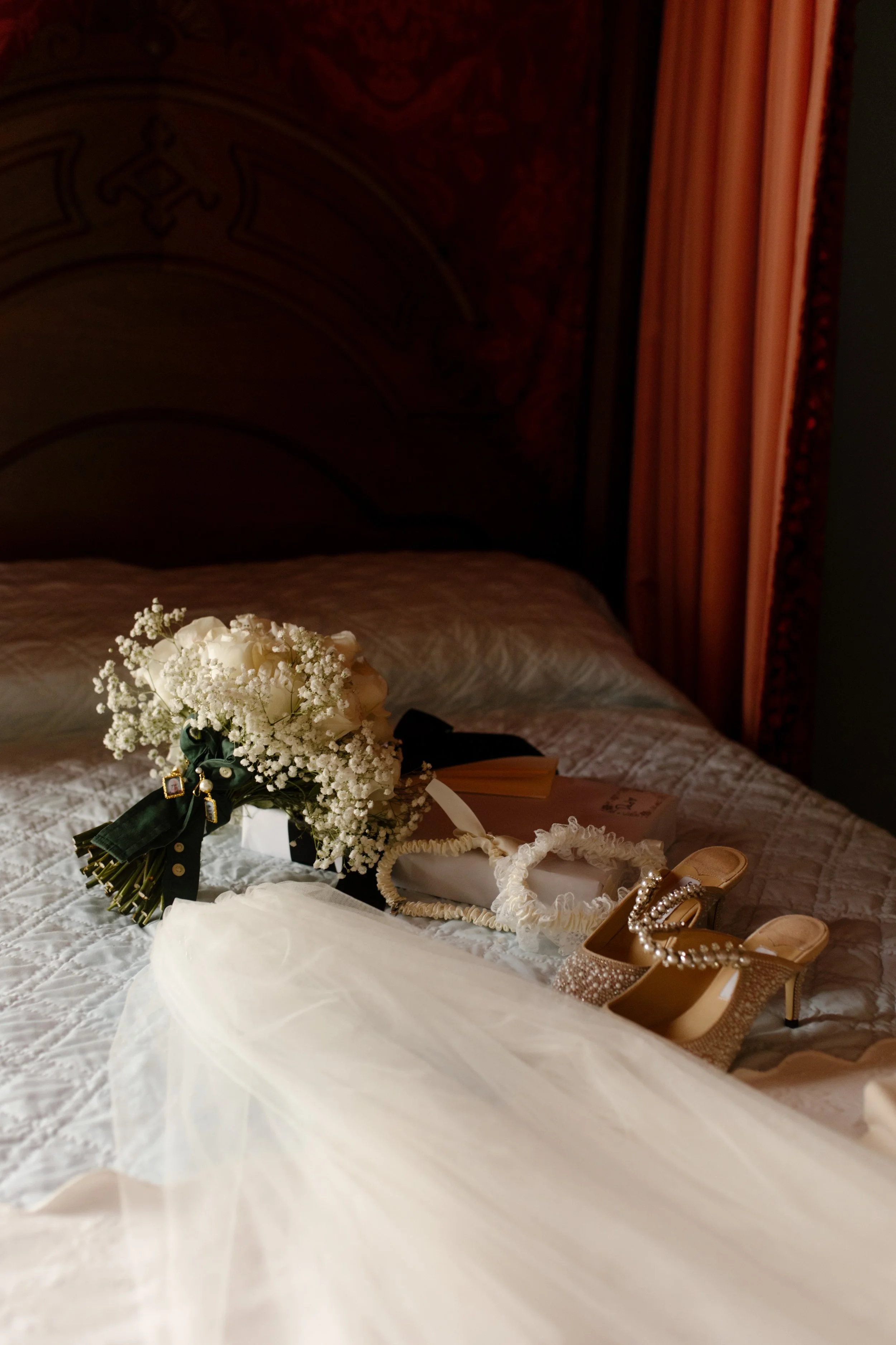 The bride’s bouquet, veil, sparkling heels, and garter rest on a vintage bedspread, captured in that quiet in-between before it all begins.