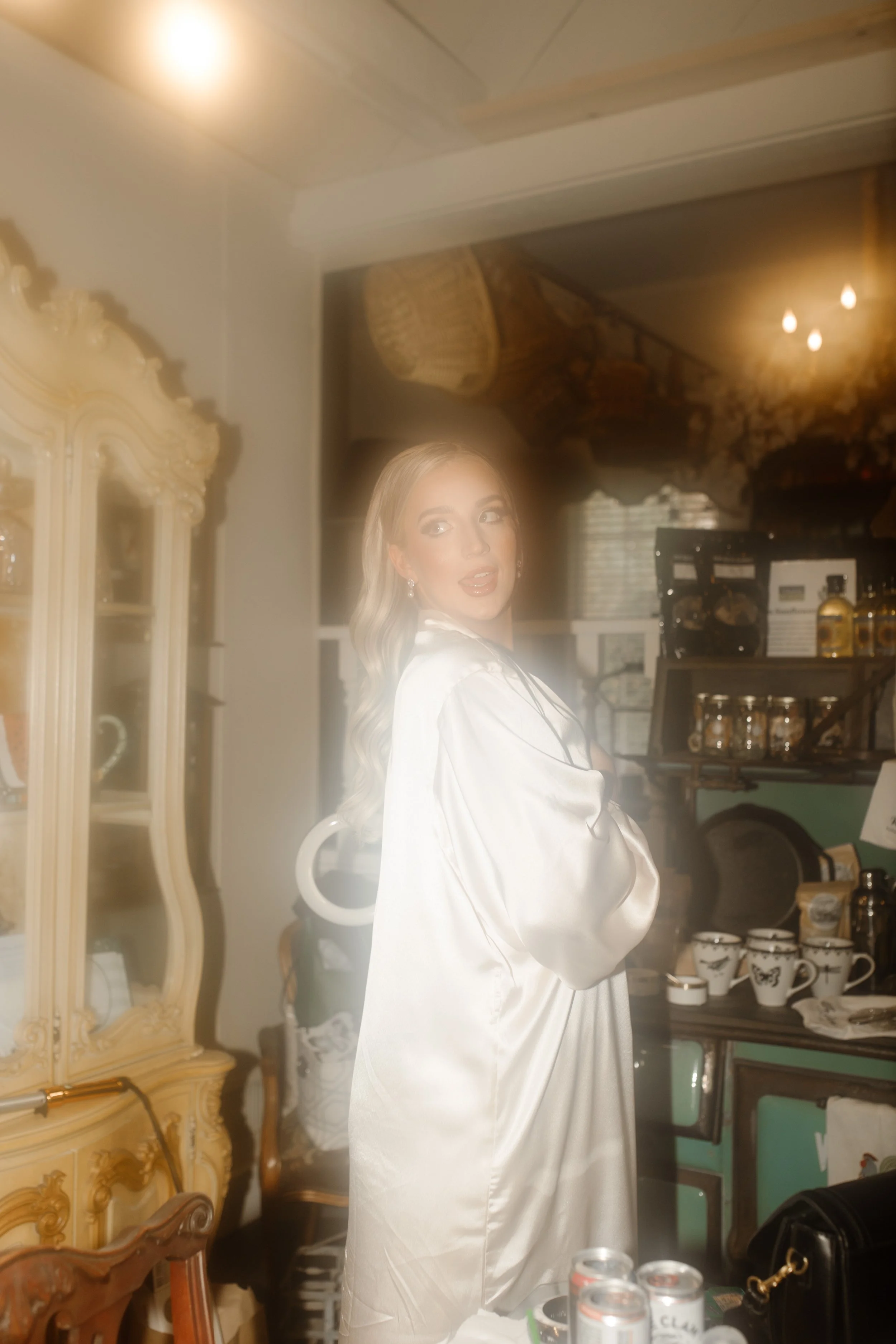 The bride glances back mid-laugh, wrapped in a satin robe and soft morning light, surrounded by vintage details at her Arkansas wedding.