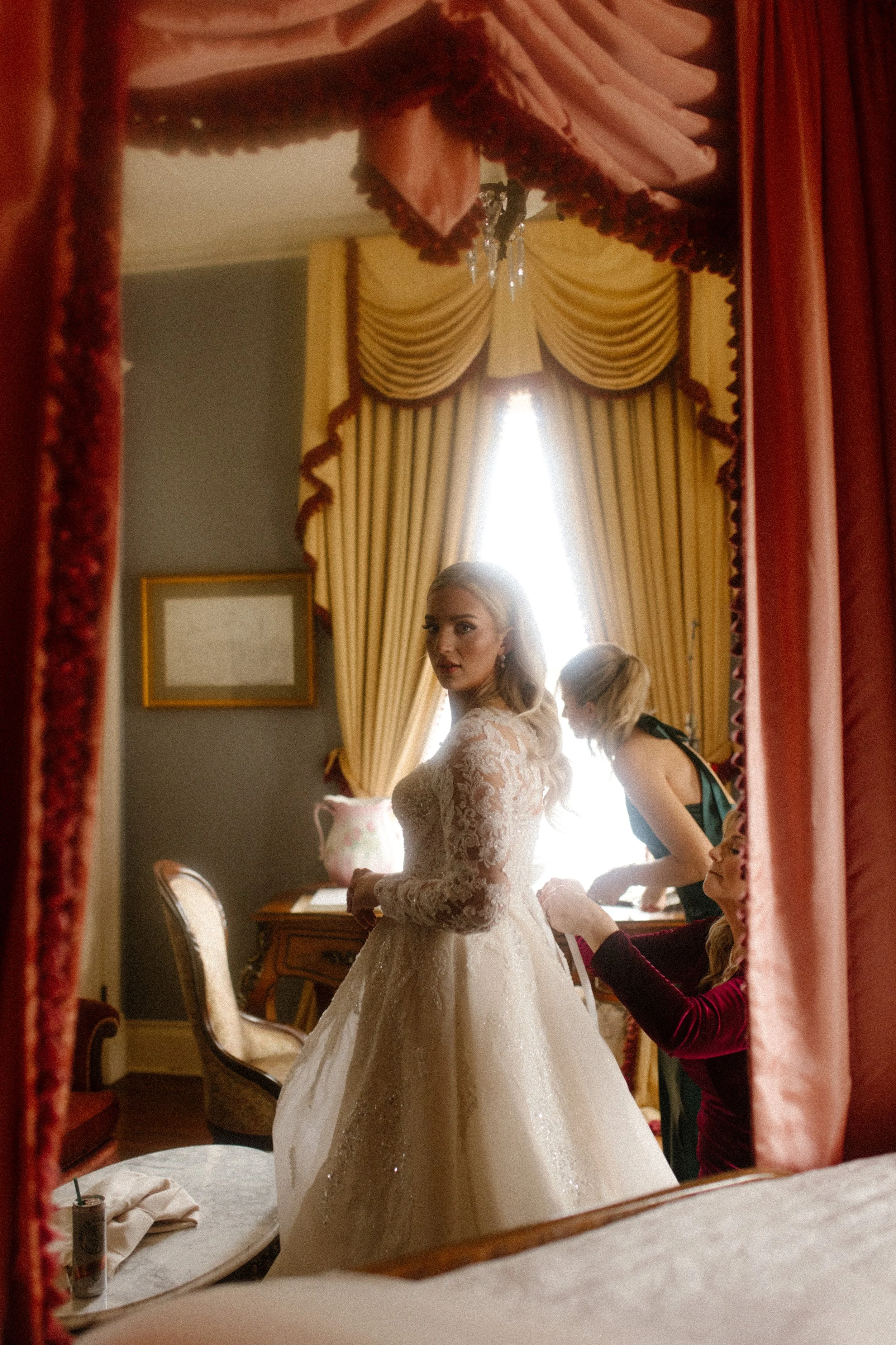 Mother of the bride helps tie the back of her daughter’s gown, surrounded by rich velvet drapes and golden light before the Arkansas wedding begins.