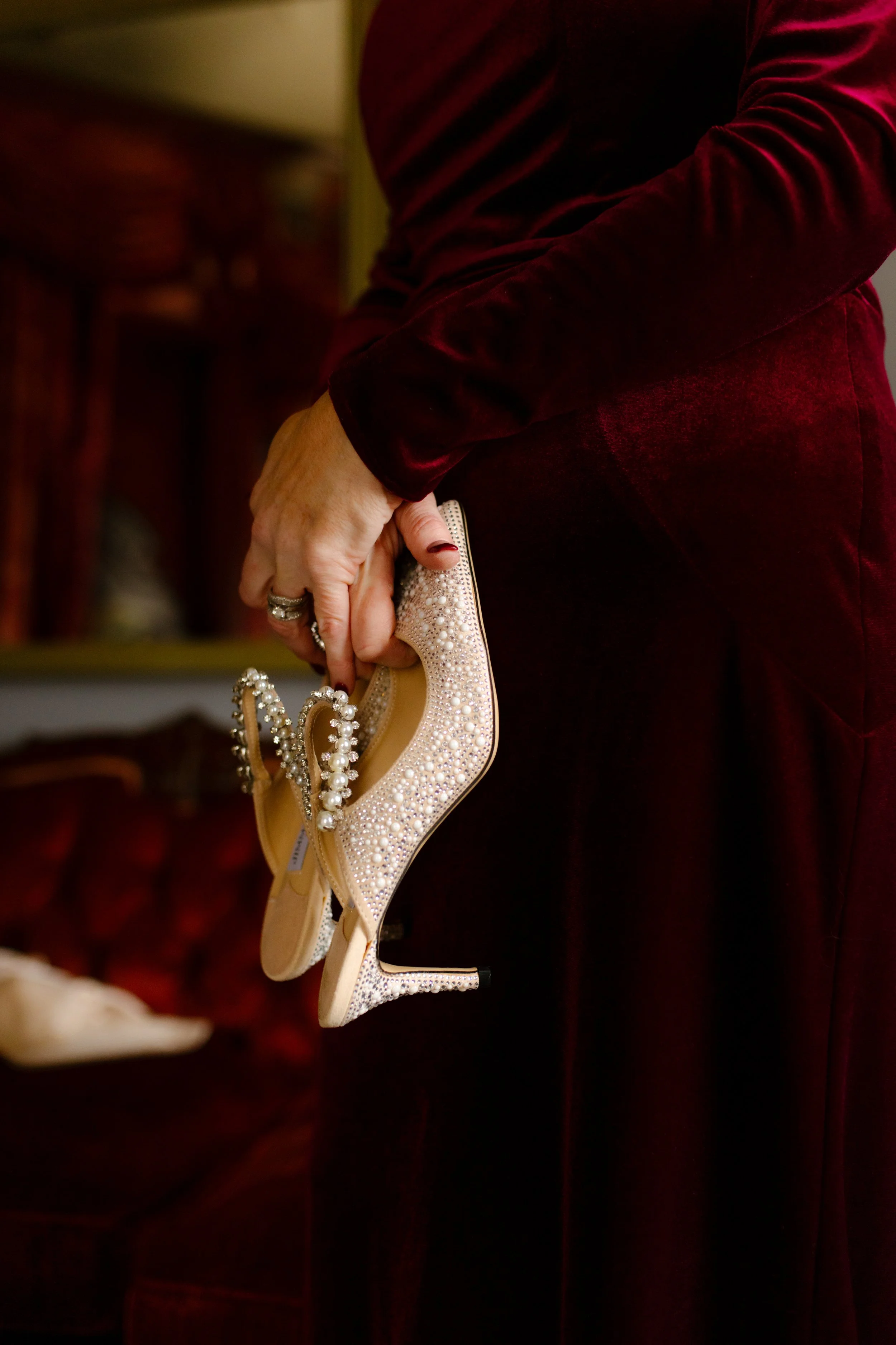 A close-up of a woman in a deep red dress holding the bride’s pearl-studded heels before the ceremony begins.