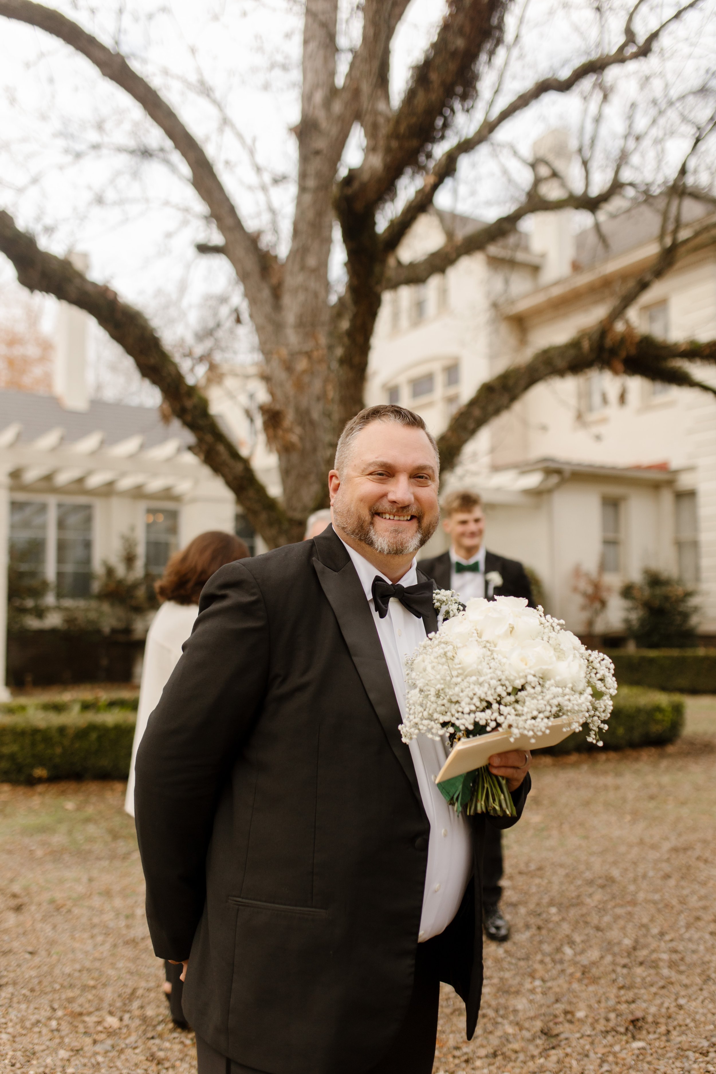 Father of the bride stands proudly with bouquet in hand, smiling wide just before walking his daughter down the aisle.