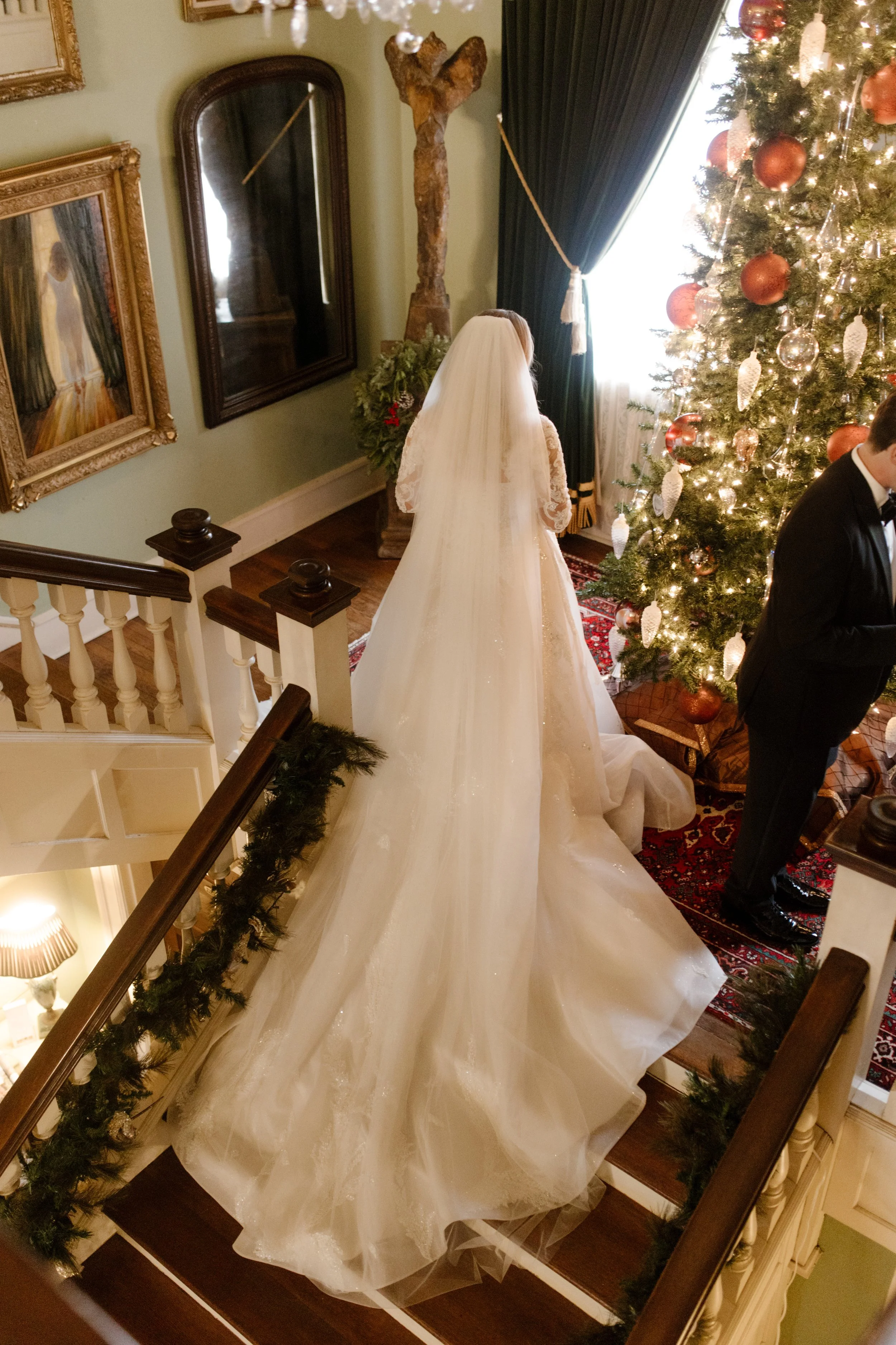 With her gown flowing behind her, the bride descends a garland-lined staircase toward the Christmas tree and her soon-to-be husband at their Arkansas wedding.