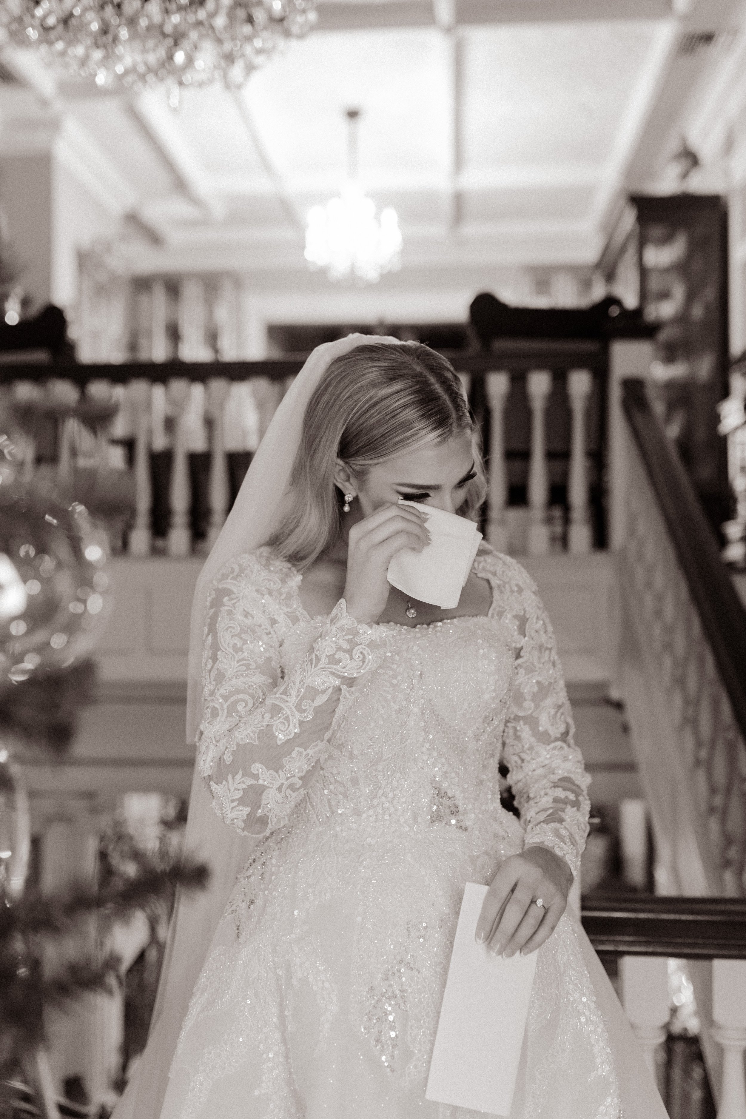 Bride stands at the foot of a grand staircase, wiping a tear as she clutches a letter, moments before the ceremony begins.