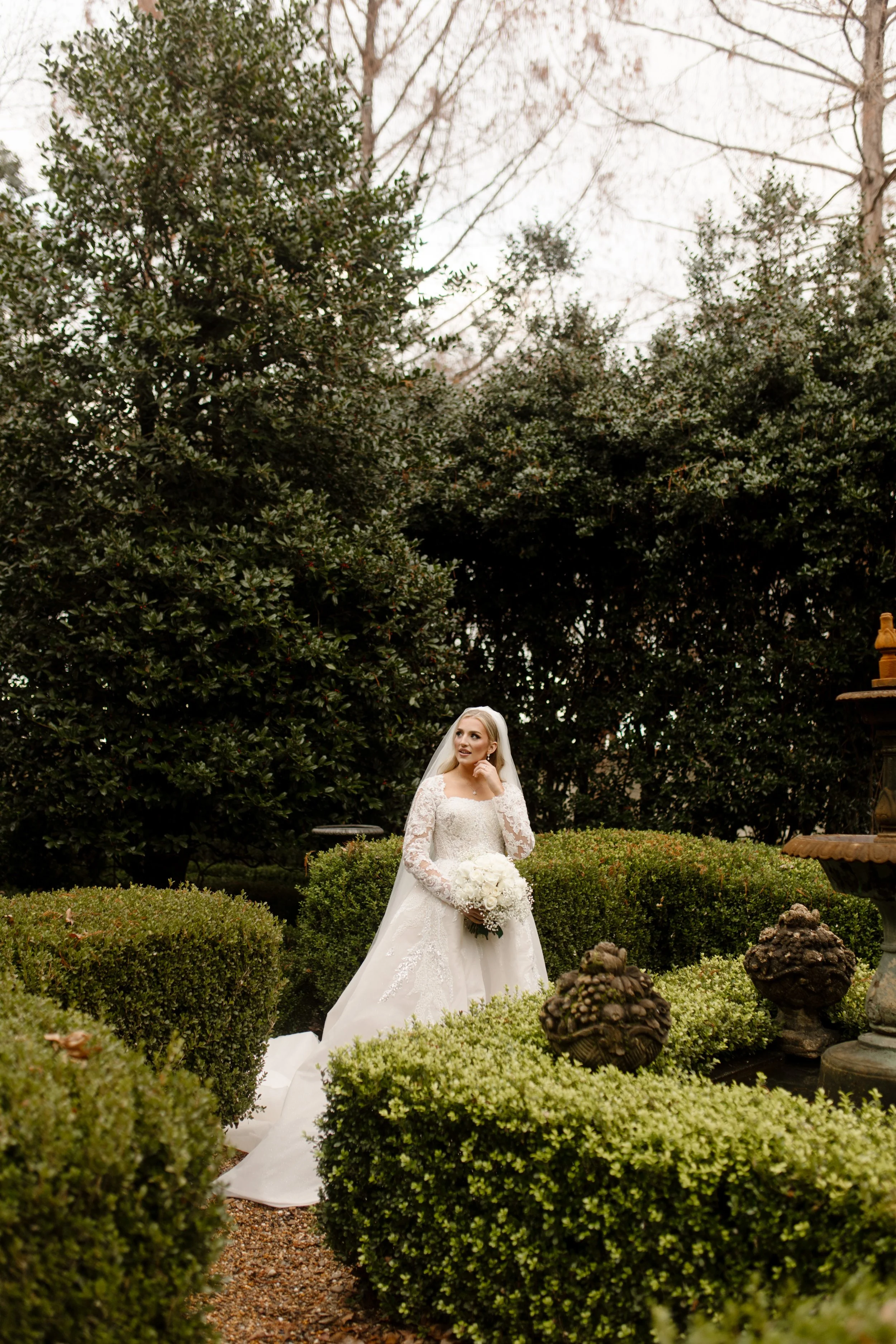 Bride stands quietly in a sculpted garden path, holding a soft bouquet of white florals, her veil trailing behind her at this Arkansas wedding.