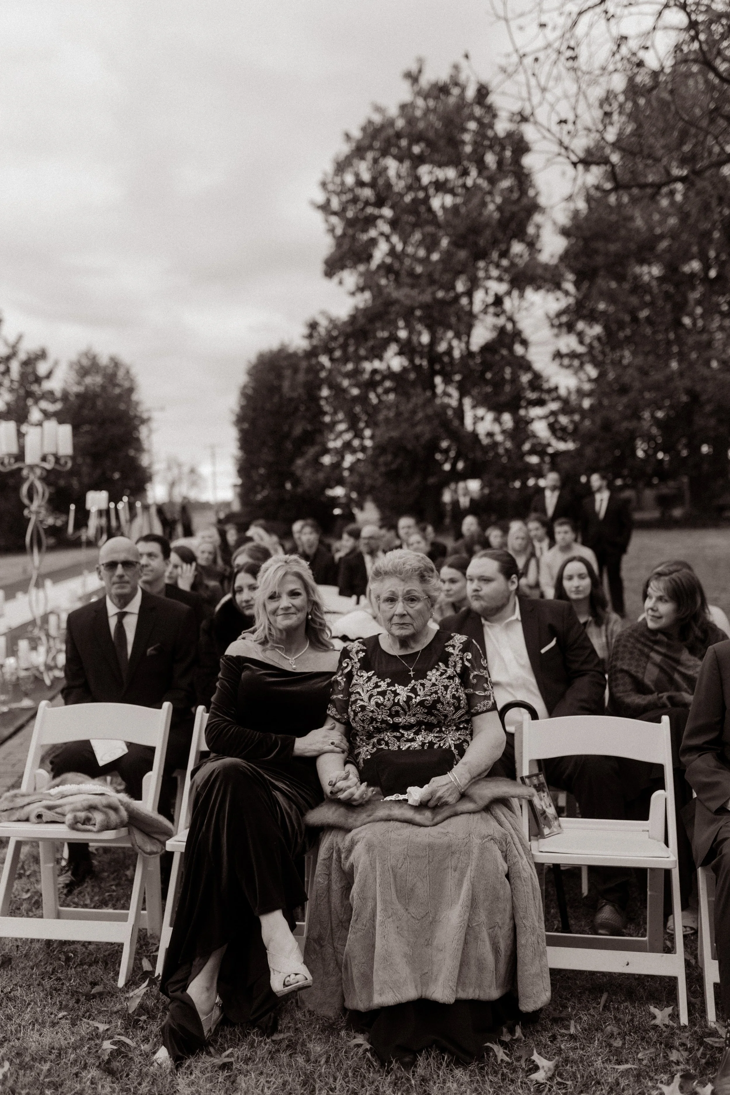 Two women—likely mother and grandmother—hold hands in the front row before the ceremony, surrounded by bundled-up guests beneath a moody sky.